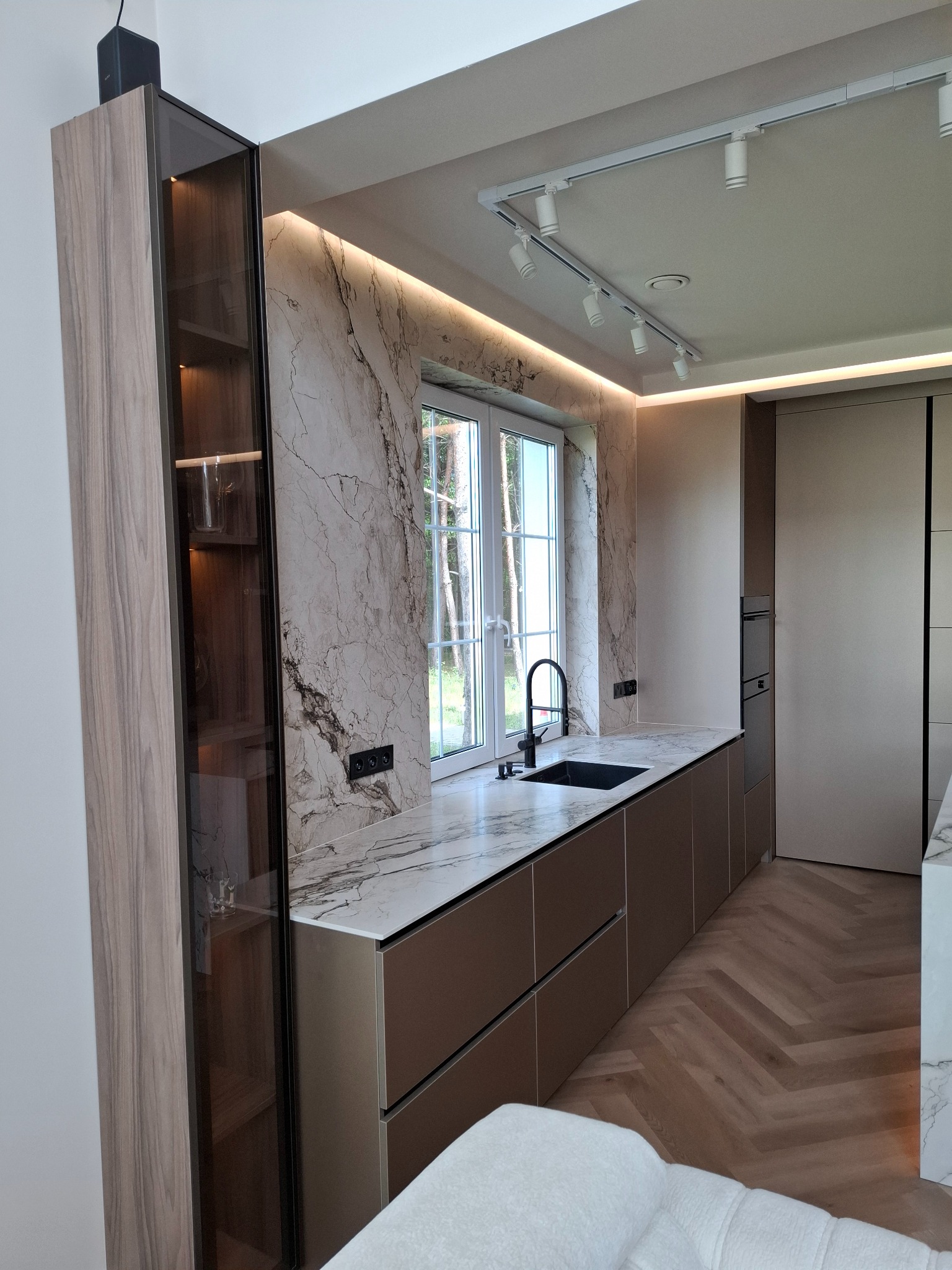 Nowoczesna kuchnia z marmurowym blatem i ścianą. Szafki w odcieniach brązu, oświetlenie punktowe i drewniana podłoga w jodełkę tworzą elegancką przestrzeń.