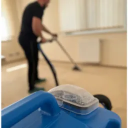 Mężczyzna w czarnym stroju czyści dywan beżową maszyną. Na pierwszym planie niebieski zbiornik z wodą i pianą. Jasne wnętrze z grzejnikiem w tle.