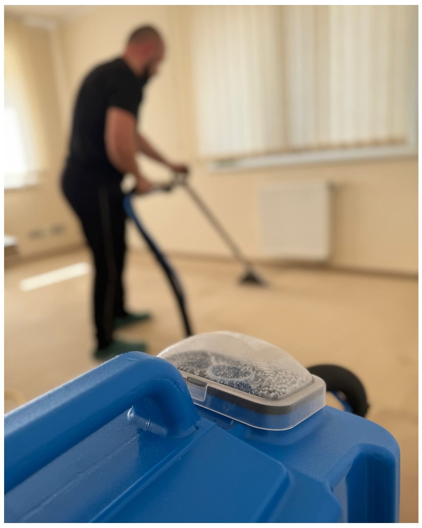 Mężczyzna w czarnym stroju czyści dywan beżową maszyną. Na pierwszym planie niebieski zbiornik z wodą i pianą. Jasne wnętrze z grzejnikiem w tle.