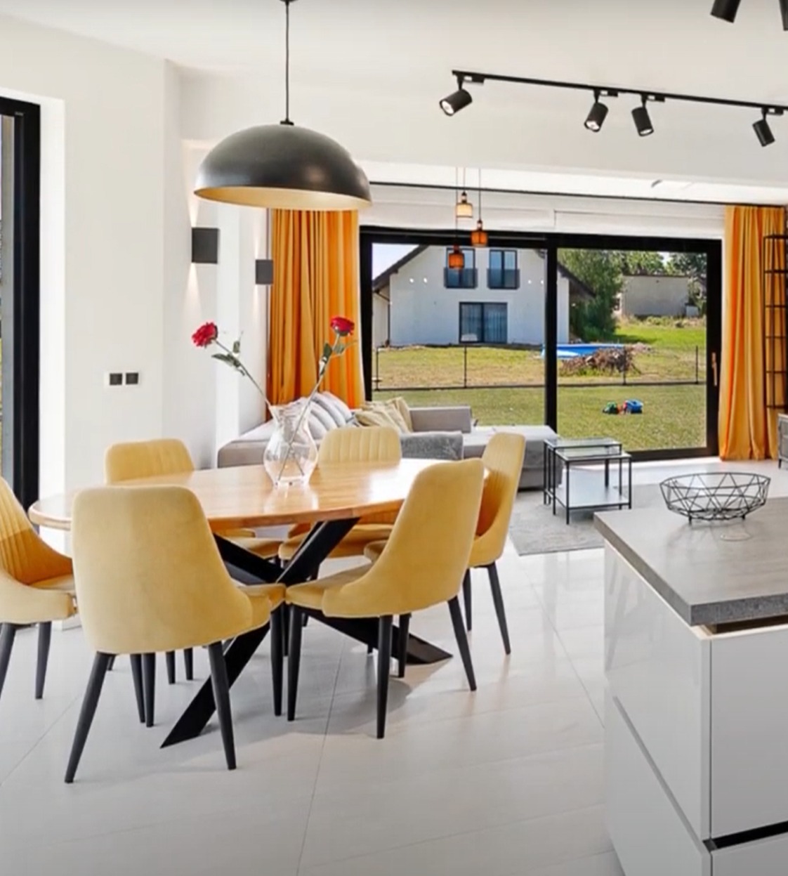 Jasne wnętrze jadalni z drewnianym stołem i żółtymi krzesłami. Za oknem widok na ogród z basenem. Nowoczesne oświetlenie punktowe.