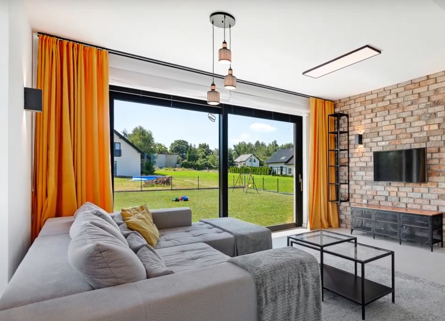 Jasny salon z ceglaną ścianą, szarą sofą i widokiem na ogród przez duże okno. Żółte zasłony i lampa wisząca dodają ciepła wnętrzu. Widoczne meble i dekoracje.