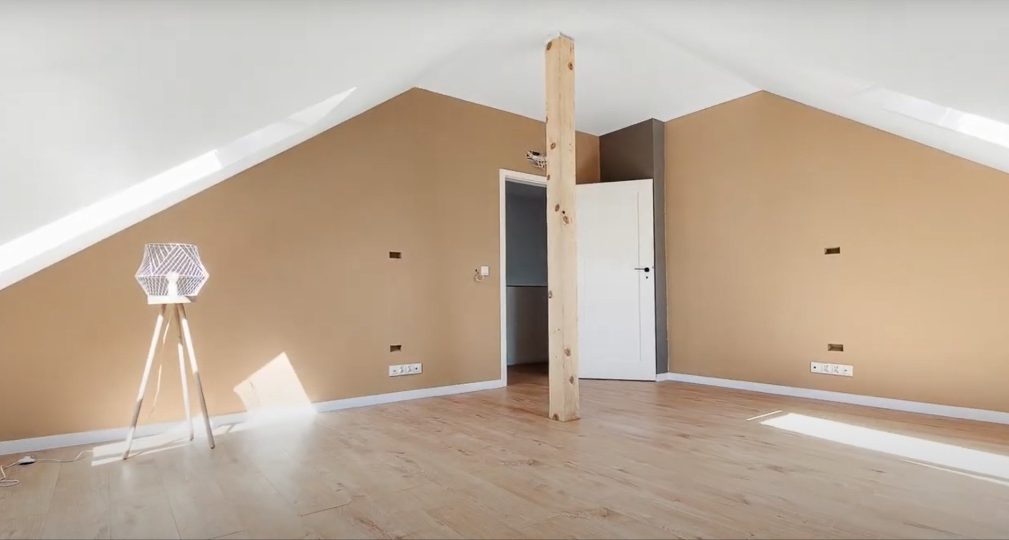 Jasne poddasze z drewnianą podłogą, beżowymi ścianami i białym sufitem. Widoczna lampa na trójnogu, drzwi i drewniany słup. Przygotowanie do wykończenia.