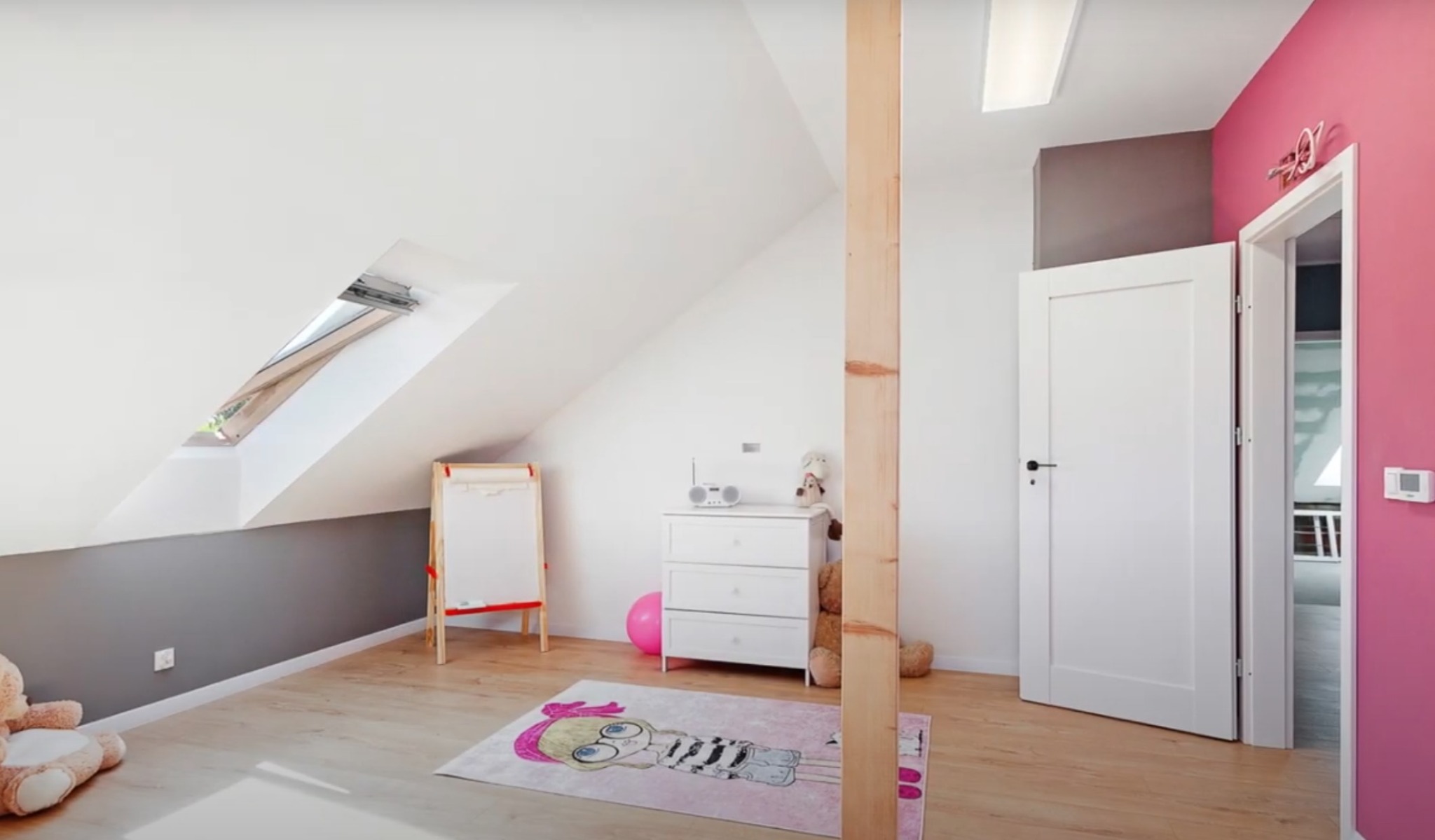 Pokój dziecięcy na poddaszu z oknem dachowym, pomalowany na biało i szaro, z różowym akcentem na ścianie przy drzwiach. Widoczne meble i zabawki.