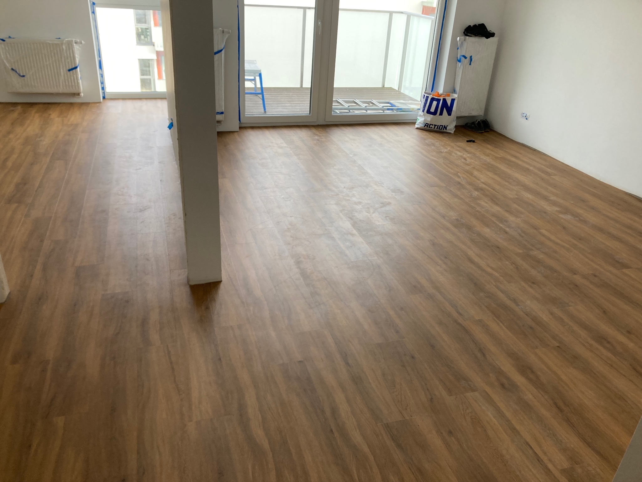 Ułożone panele podłogowe w pokoju z wyjściem na balkon. Grzejnik i framugi okien zabezpieczone folią. Widoczny worek z zakupami.