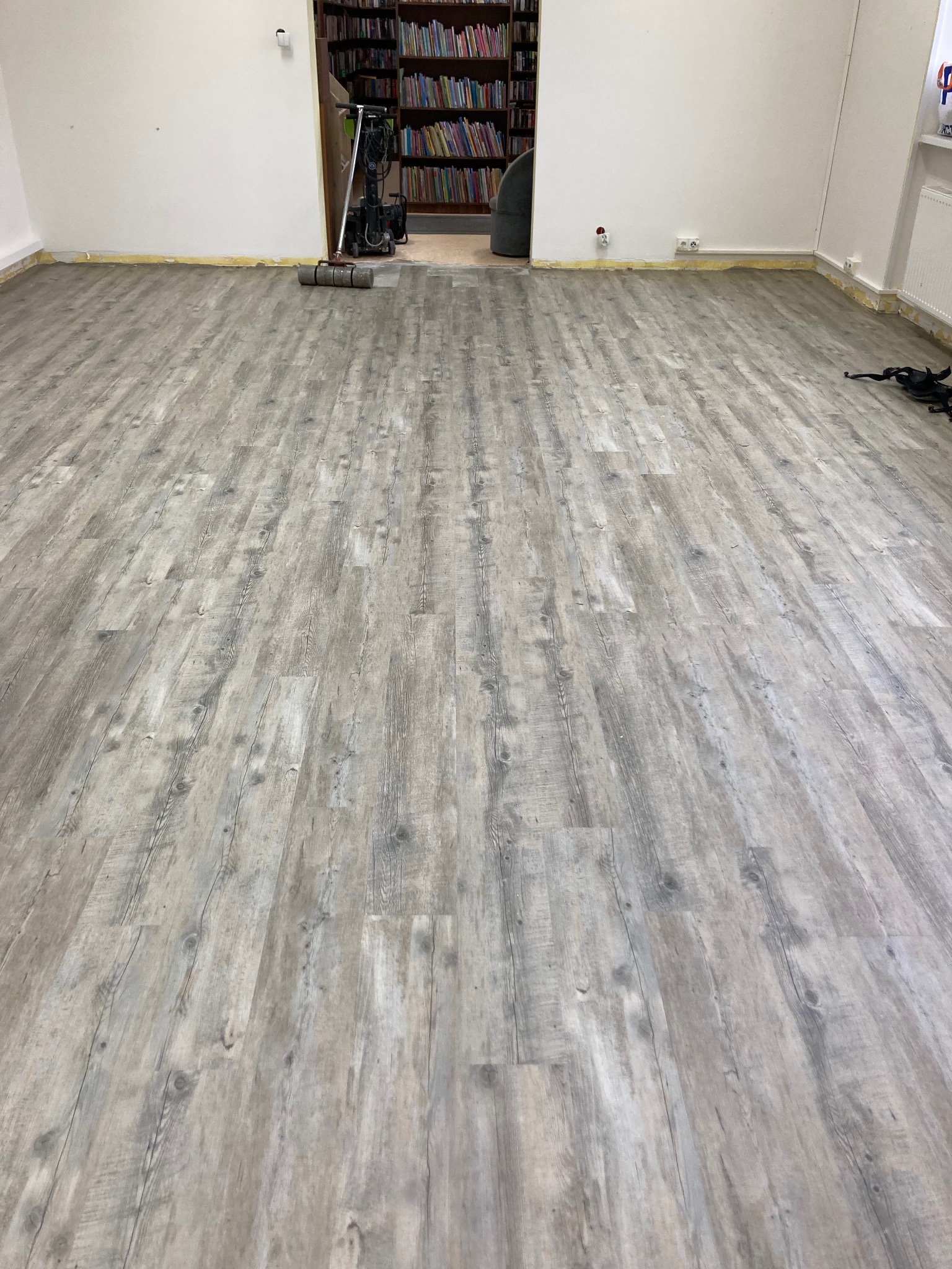 Jasnoszara podłoga z paneli imitujących drewno, ułożona w pokoju z białymi ścianami. W tle widoczne regały z książkami i sprzęt do sprzątania.