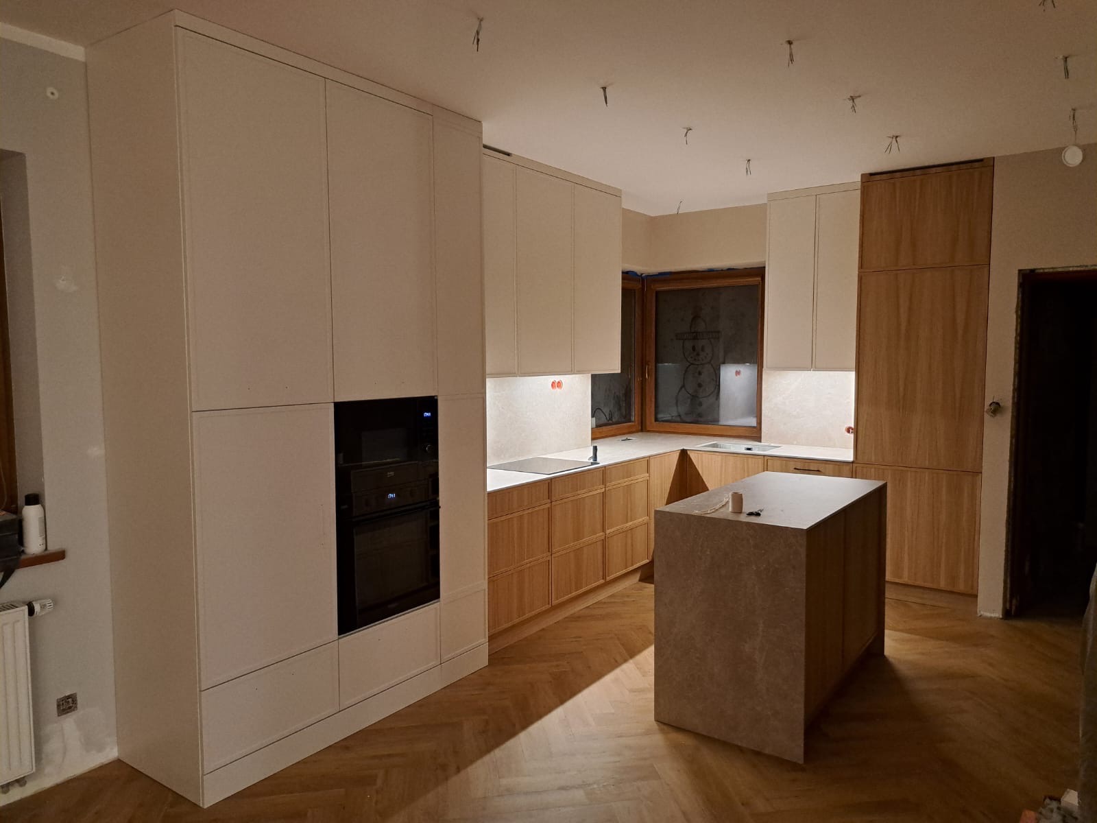 Nowoczesna kuchnia z wyspą, jasne fronty szafek i drewniane wykończenia, parkiet w jodełkę. Widoczne niedokończone prace wykończeniowe na suficie.