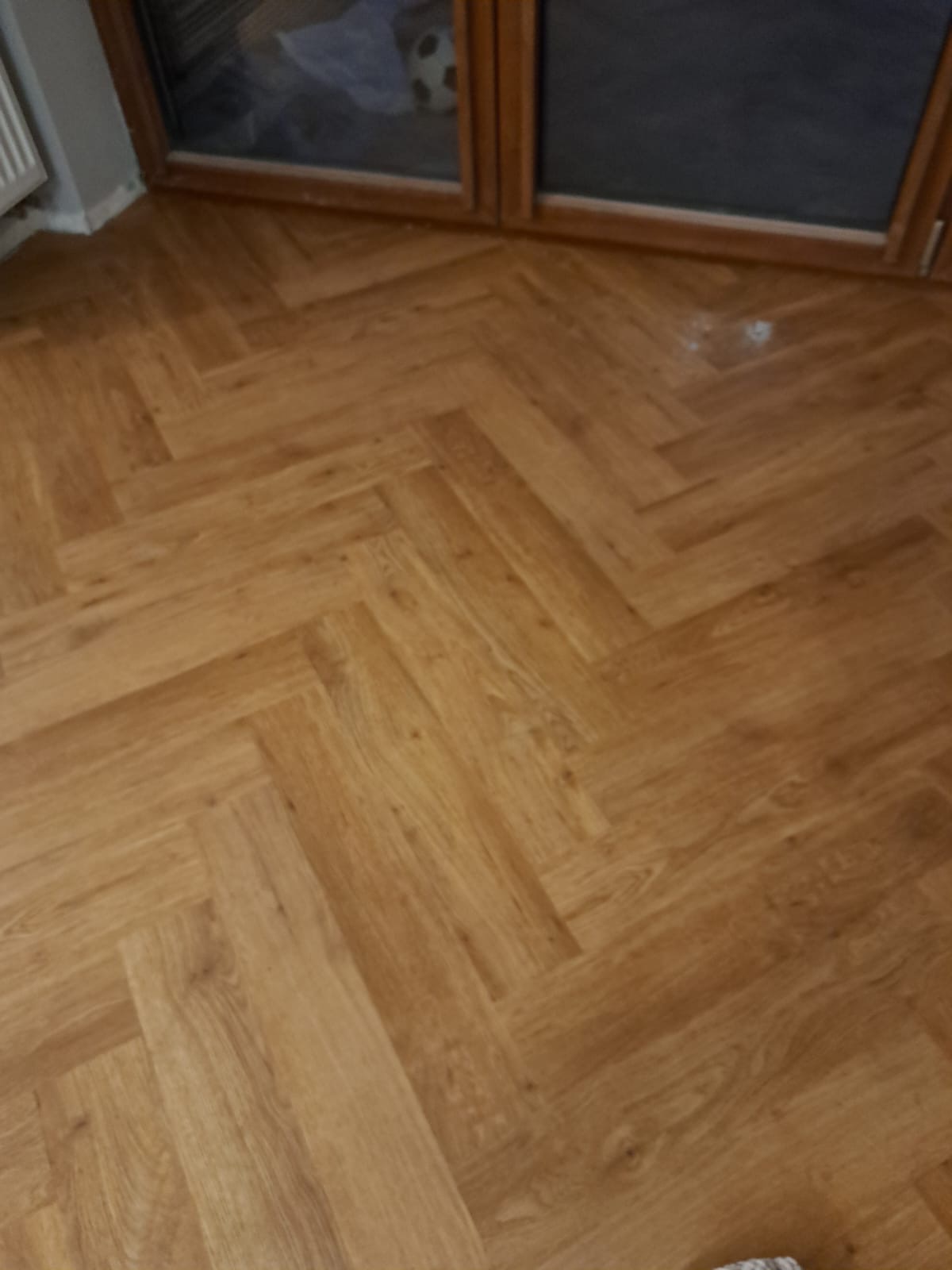 Ułożona podłoga z paneli w jodełkę, widoczna przy oknie. Jasne, drewnopodobne panele tworzą wzór na podłodze. Fragment grzejnika i okna w tle.