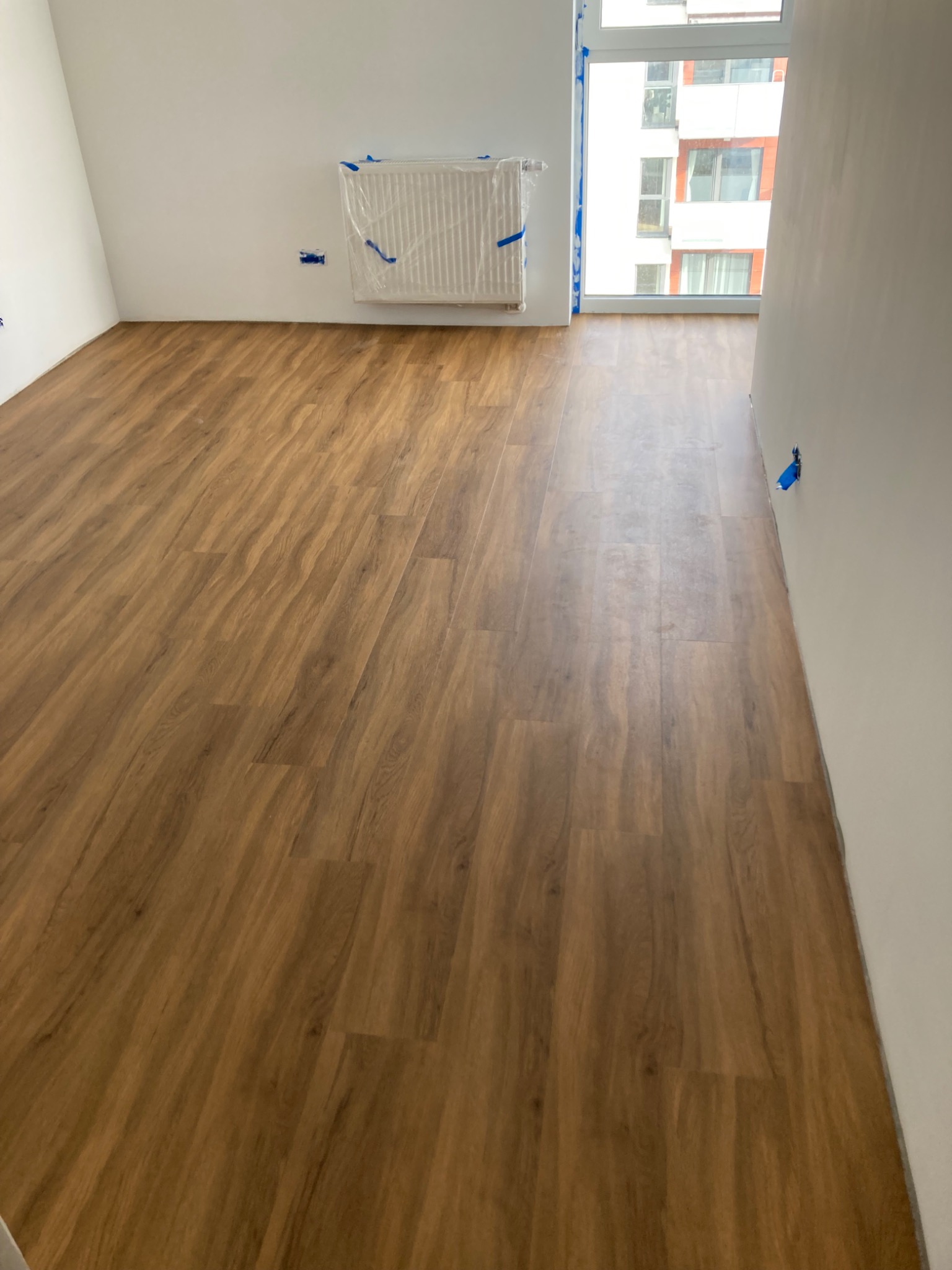 Jasnobrązowe panele podłogowe ułożone w pustym pokoju z oknem. Grzejnik zabezpieczony folią malarską. Świeżo wykończone ściany w neutralnym odcieniu bieli.