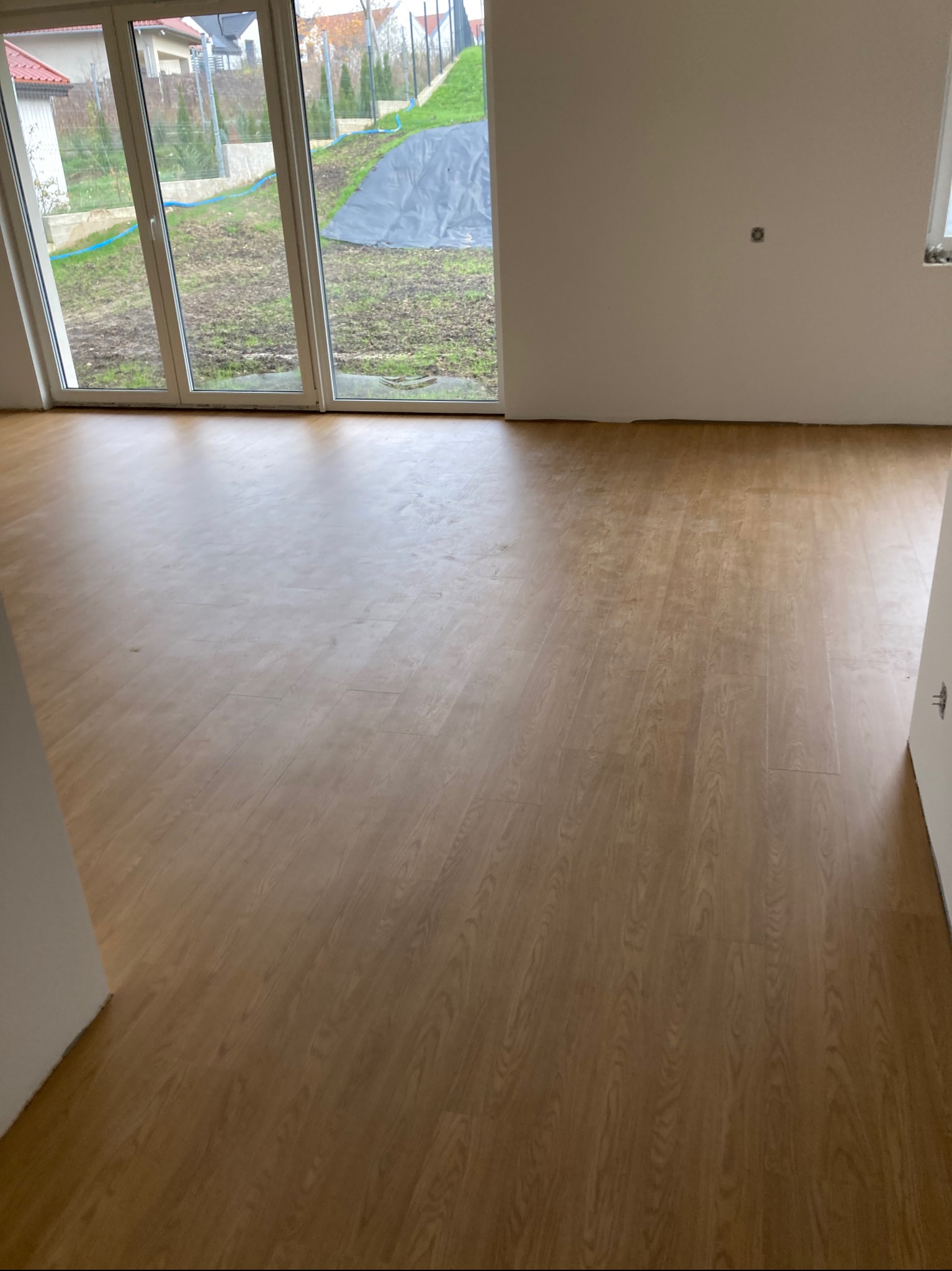 Jasne panele podłogowe ułożone w nowym, pustym pokoju z dużym oknem tarasowym wychodzącym na ogród. Widoczny wzór drewna i fuga paneli.