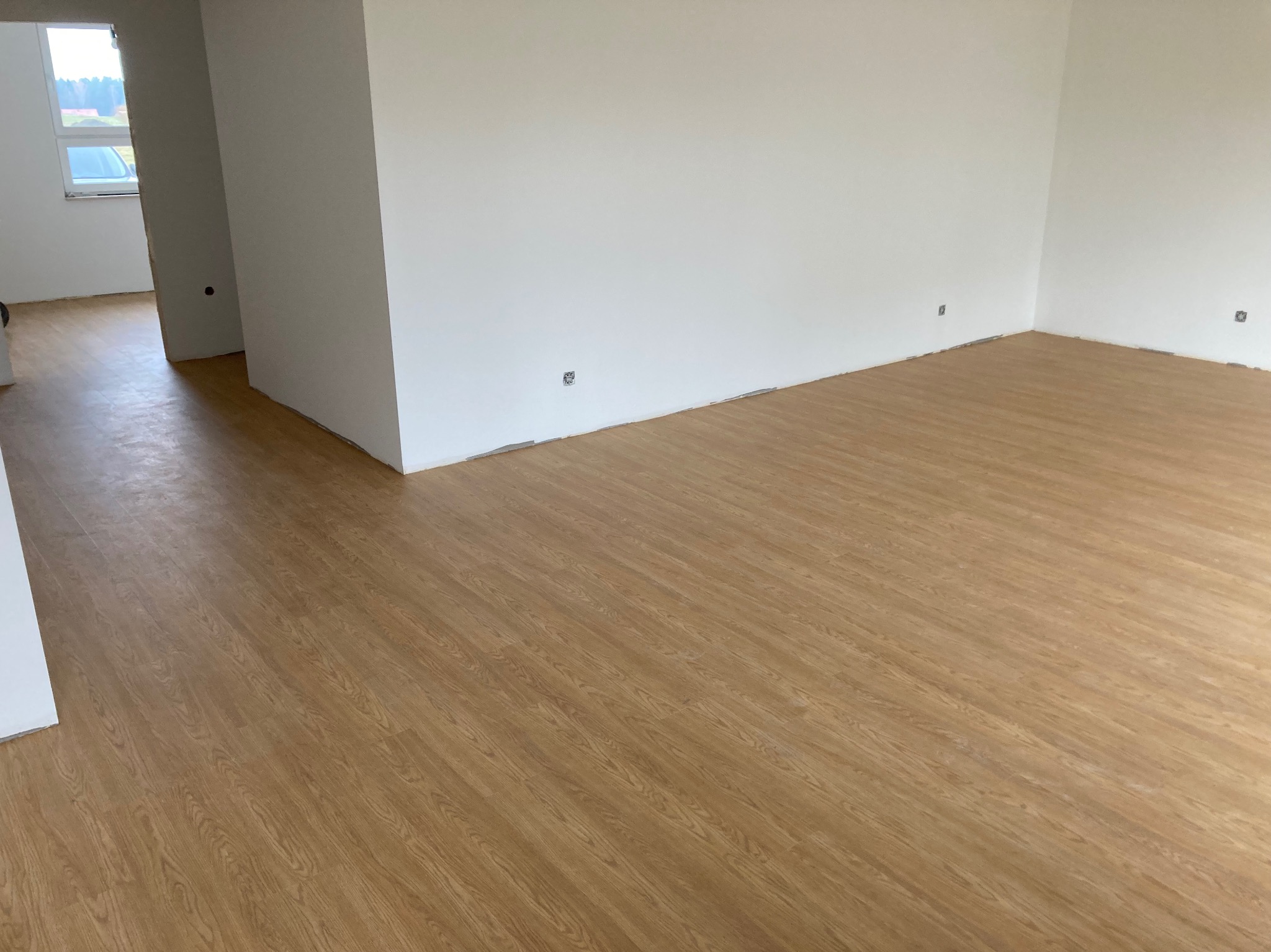 Jasne panele podłogowe w nowym, pustym pokoju z białymi ścianami i widocznymi puszkami elektrycznymi. Widok na sąsiednie pomieszczenie.