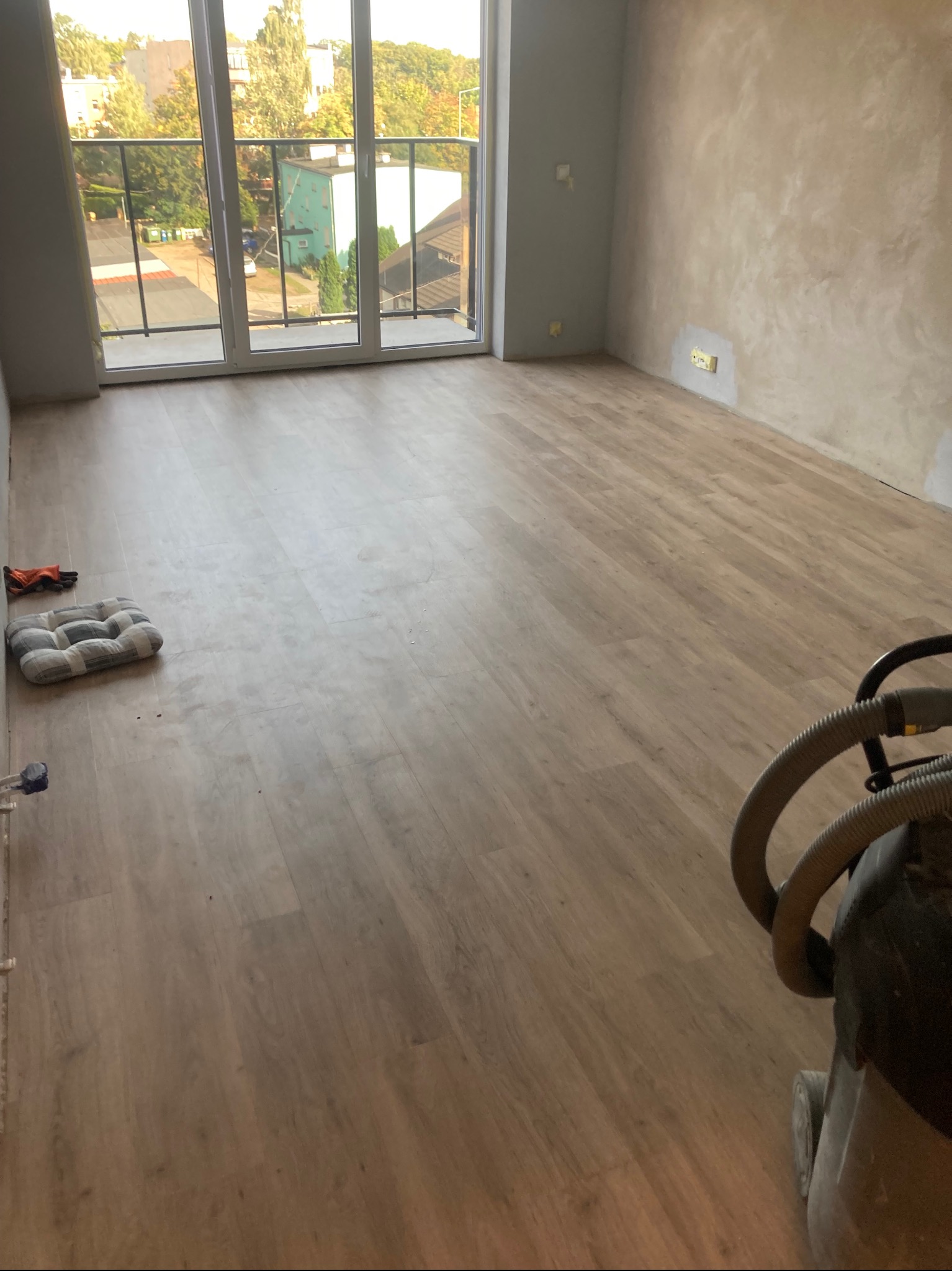 Jasne panele podłogowe w pokoju z balkonem, widoczne narzędzia pracy i poduszka. Ściany częściowo wykończone, przygotowane do malowania. Widok z okna na zieleń.