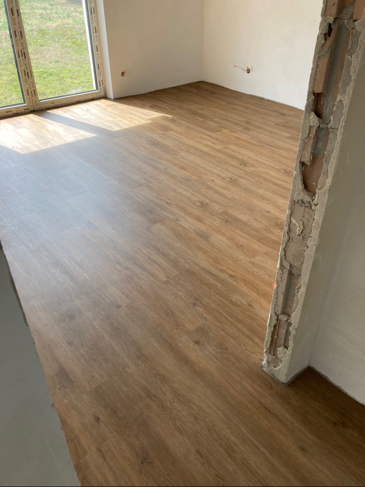 Jasne panele podłogowe w pokoju z dużym oknem i surowym wykończeniem ściany. Widoczne światło słoneczne padające na podłogę.