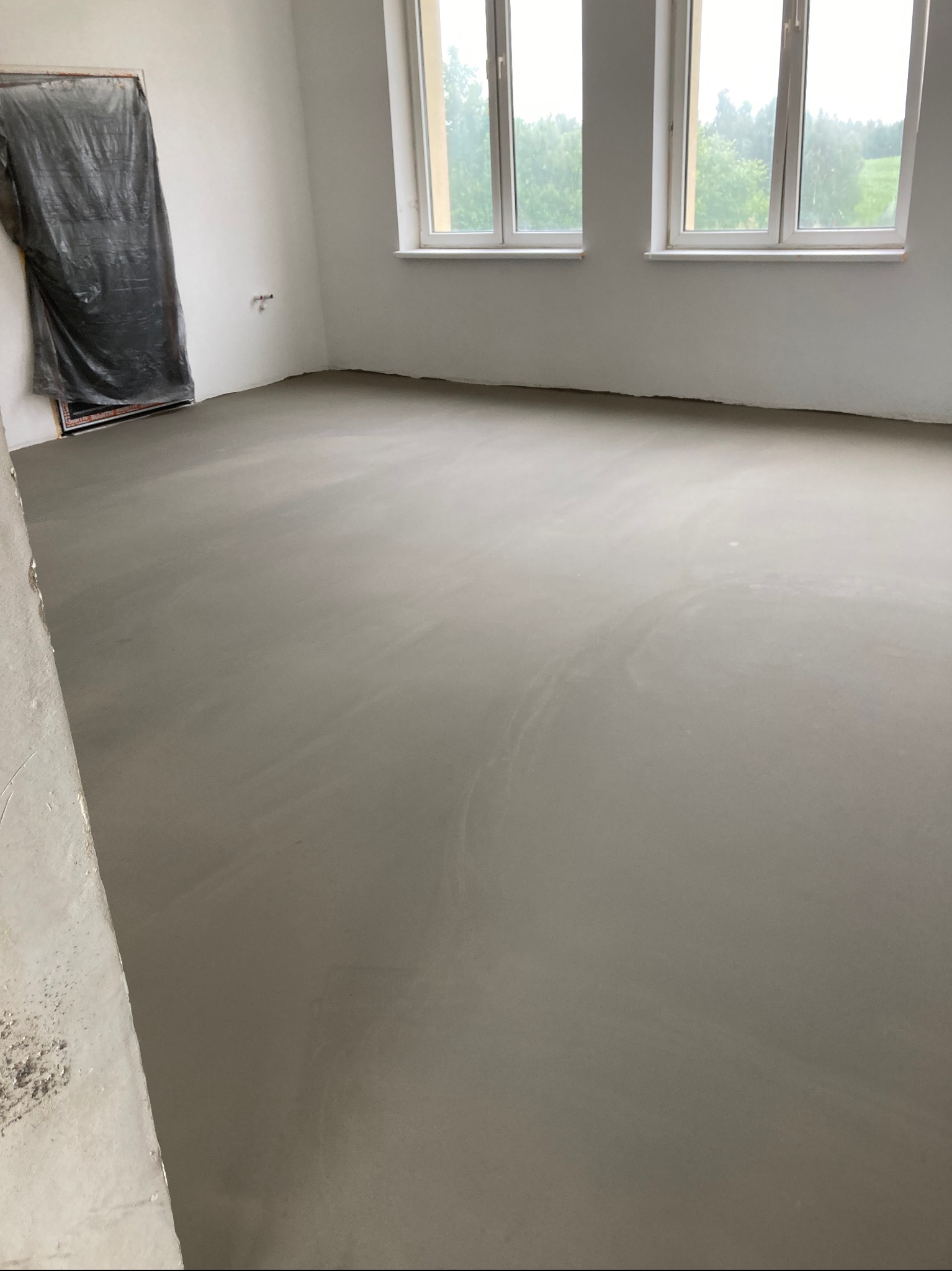 Wylana, gładka posadzka samopoziomująca w jasnym pomieszczeniu z dwoma oknami. Zabezpieczone drzwi folią malarską. Widok na zieleń za oknem.