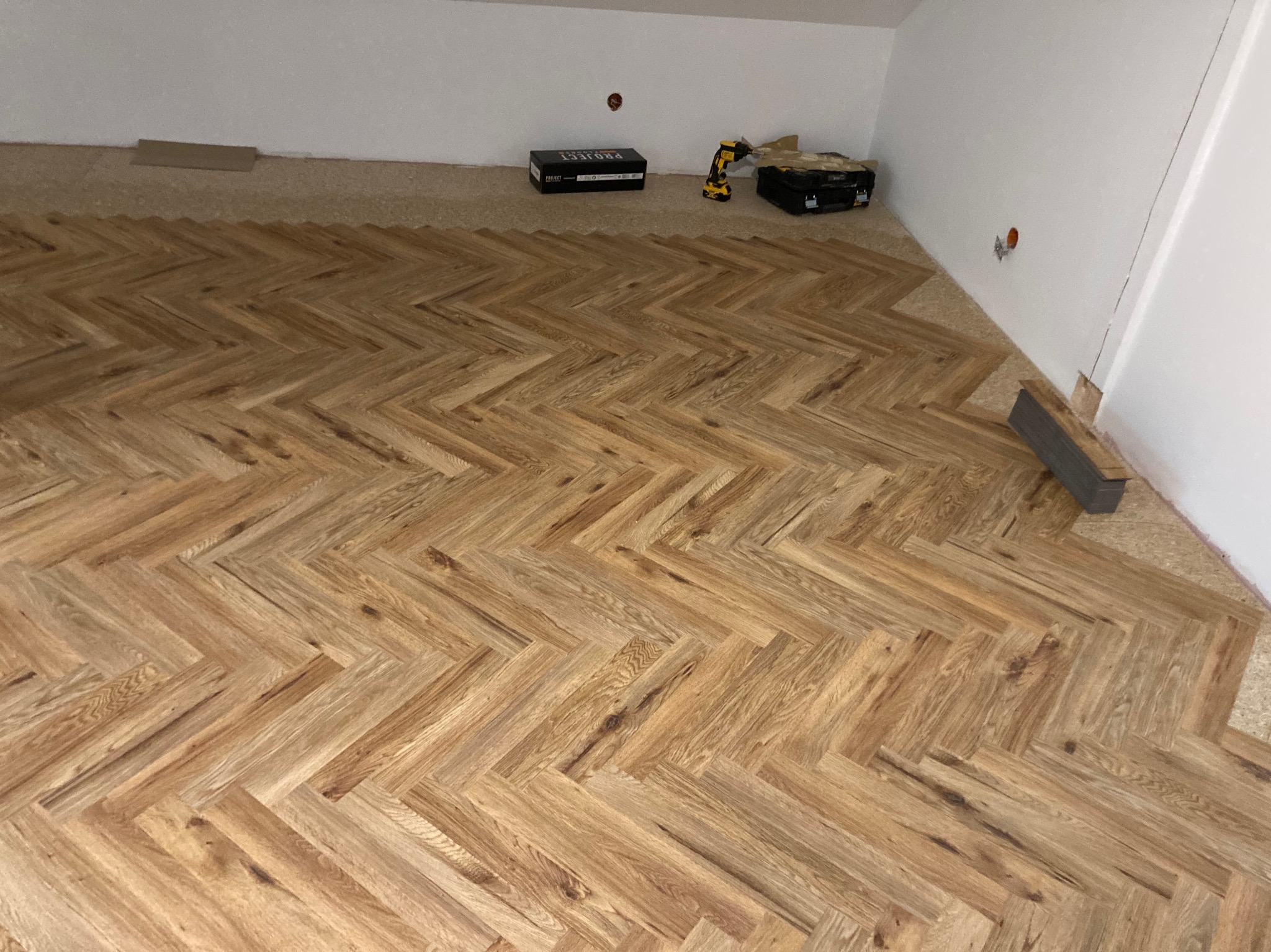 Ułożony parkiet w jodełkę, narzędzia pracy w tle. Widoczny fragment podkładu i niedokończone krawędzie przy ścianach. Jasne drewno, białe ściany.