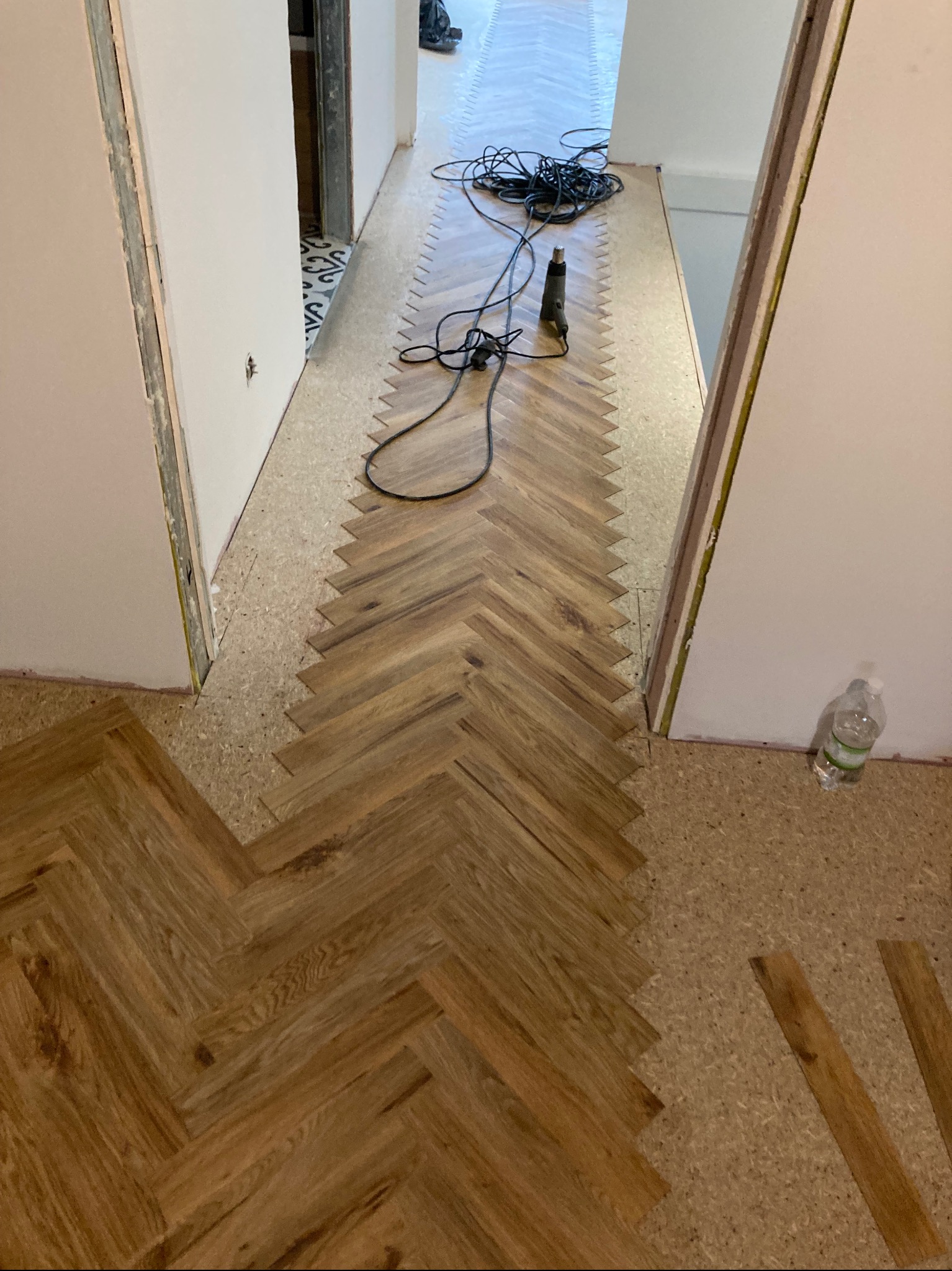 Fragment układania dębowej podłogi w jodełkę, widoczne narzędzia i podkład korkowy. Częściowo wykończone przejście między pokojami.