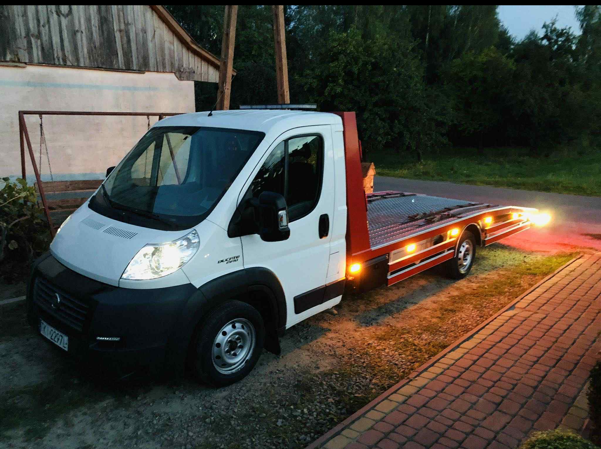 Biała autolaweta Fiat Ducato z czerwoną platformą, oświetlona, stoi na brukowanej drodze przed budynkiem i zielenią. Widoczne światła pozycyjne i tablica rejestracyjna.