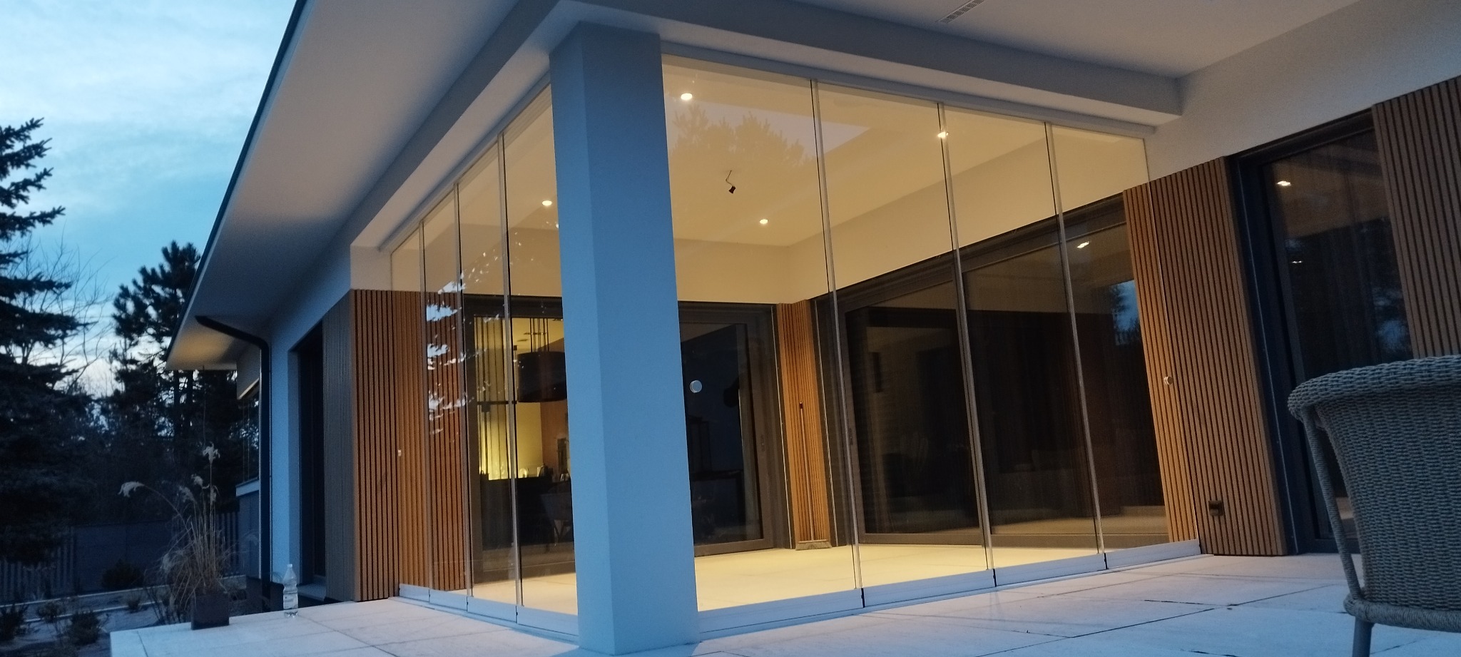 Szklana zabudowa tarasu z drewnianymi panelami, widok z zewnątrz. Nowoczesny design, wieczorne oświetlenie. Częściowo widoczne krzesło z rattanu.