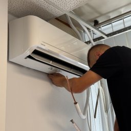 PSD SPÓŁKA Z OGRANICZONĄ ODPOWIEDZIALNOŚCIĄ - Monteraż klimatyzacji Toshiba na ścianie; widoczne rury, miernik i sufit podwieszany. Fachowiec w czarnej koszulce przy pracy.