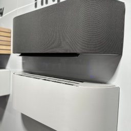 PSD SPÓŁKA Z OGRANICZONĄ ODPOWIEDZIALNOŚCIĄ - Dwa nowoczesne klimatyzatory ścienne: szary tekstylny i biały, o zaokrąglonych kształtach, zamontowane na białej ścianie. Minimalistyczny design.