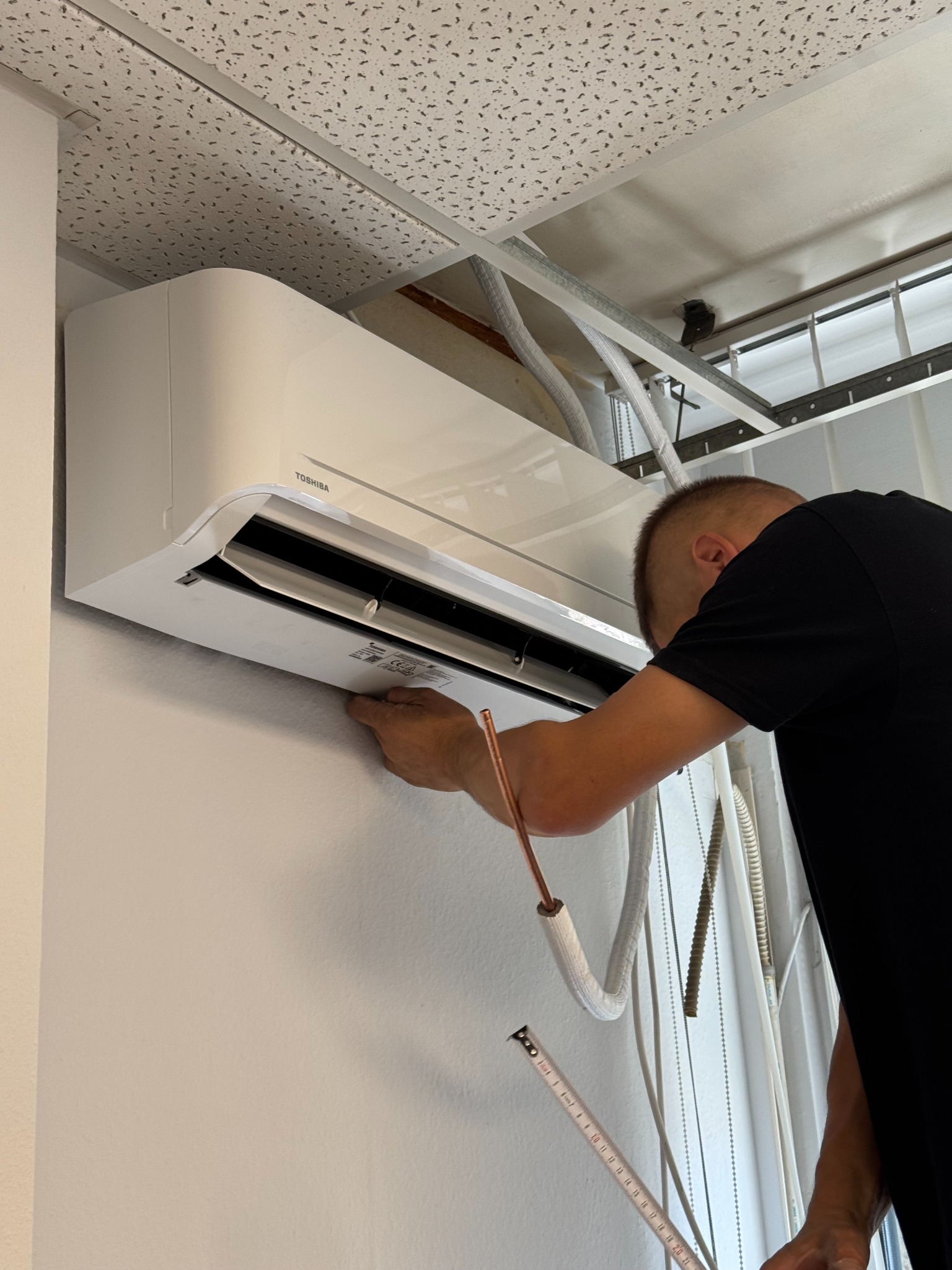 Monteraż klimatyzacji Toshiba na ścianie; widoczne rury, miernik i sufit podwieszany. Fachowiec w czarnej koszulce przy pracy.