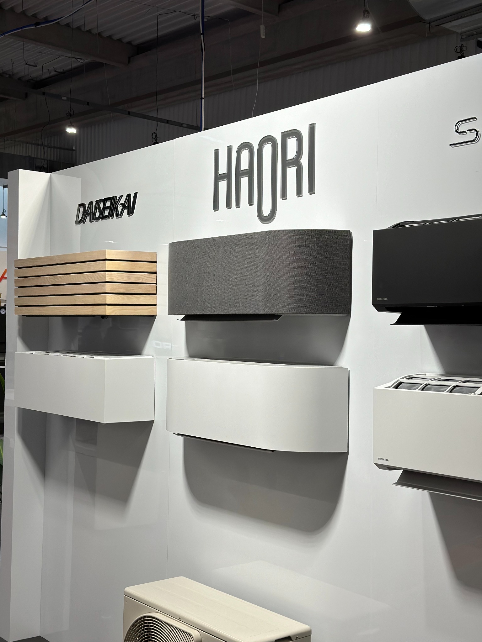 Wystawa klimatyzatorów DAISEIKAI i HAORI w różnych kolorach i wykończeniach, prezentowanych na białym tle ekspozycyjnym, z widocznym agregatem zewnętrznym.