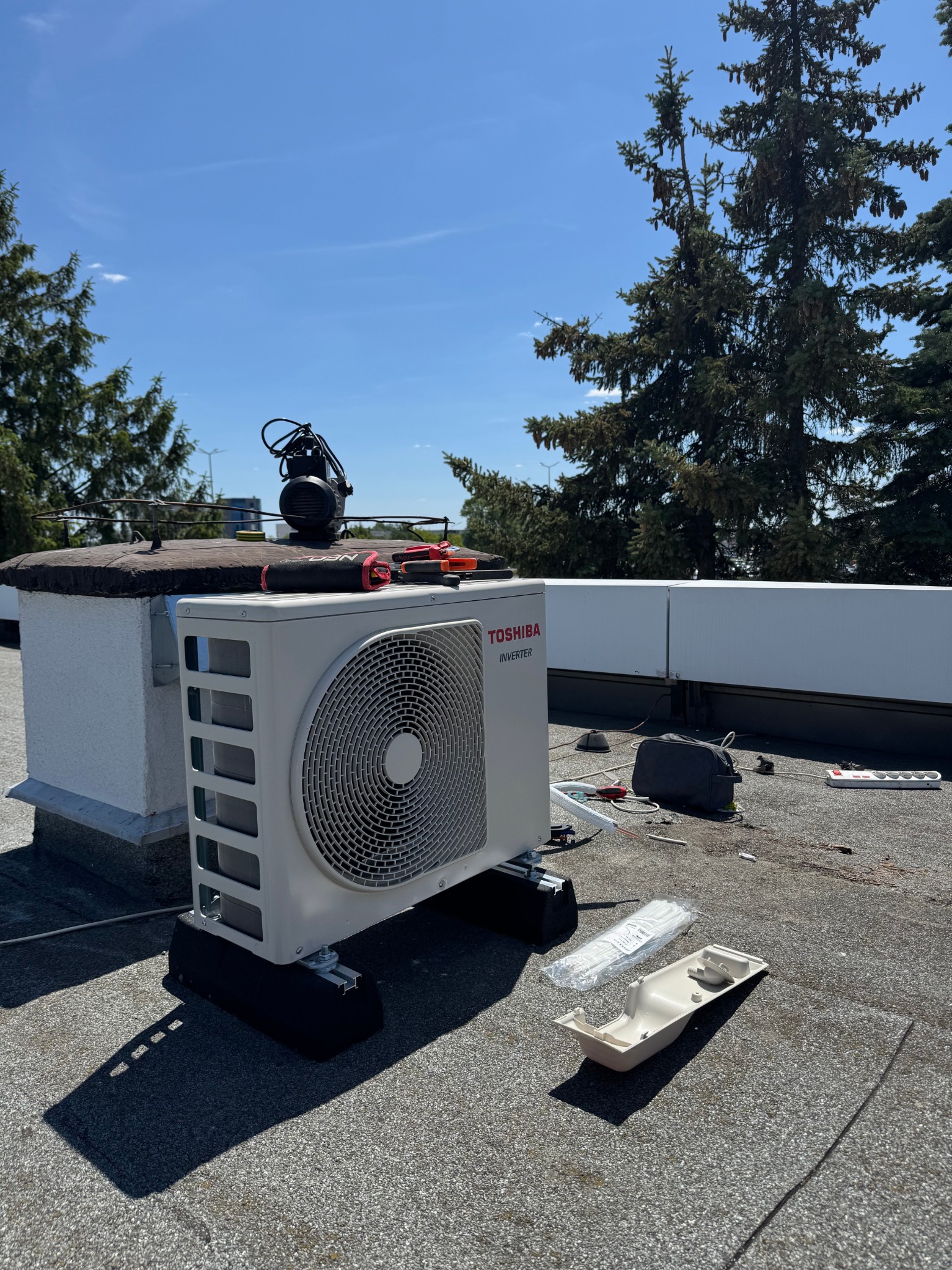 Biały klimatyzator Toshiba Inverter na dachu, narzędzia obok. Widok na błękitne niebo i drzewa iglaste. Instalacja klimatyzacji w słoneczny dzień.