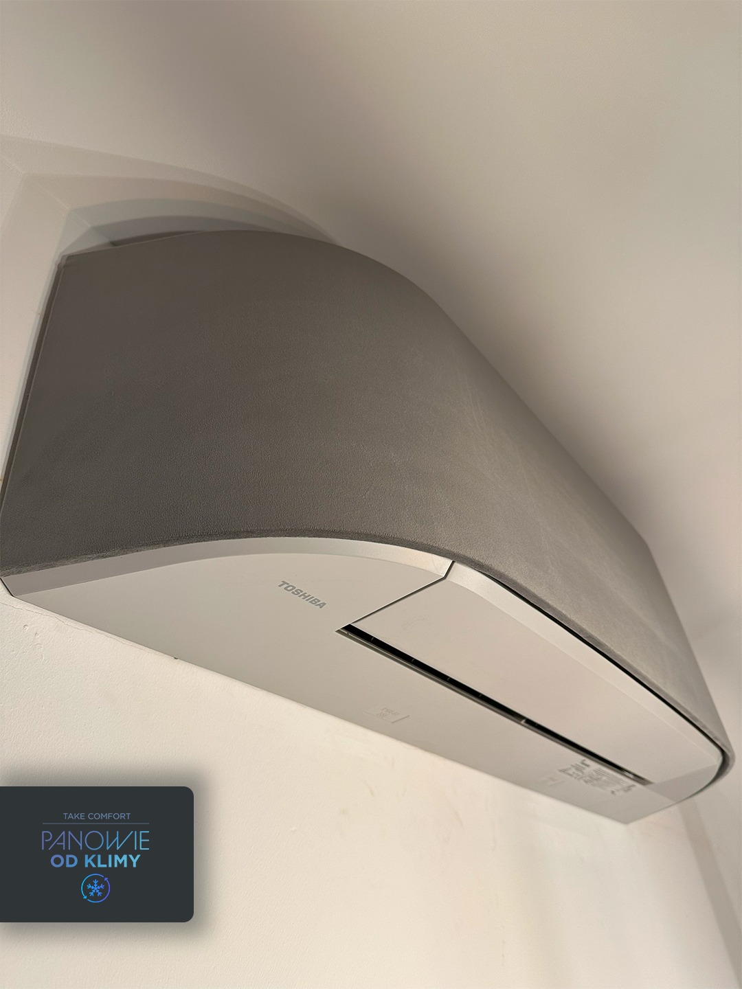 Szary klimatyzator Toshiba na ścianie, widziany z dołu, z logo firmy instalacyjnej w lewym dolnym rogu. Nowoczesny design, minimalistyczne wnętrze.