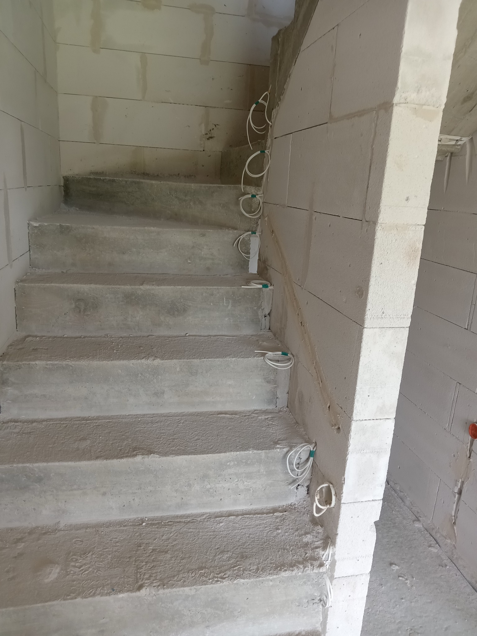 Surowe betonowe schody w trakcie budowy z wystającymi przewodami elektrycznymi w ścianie z białych bloczków. Widok z dołu.