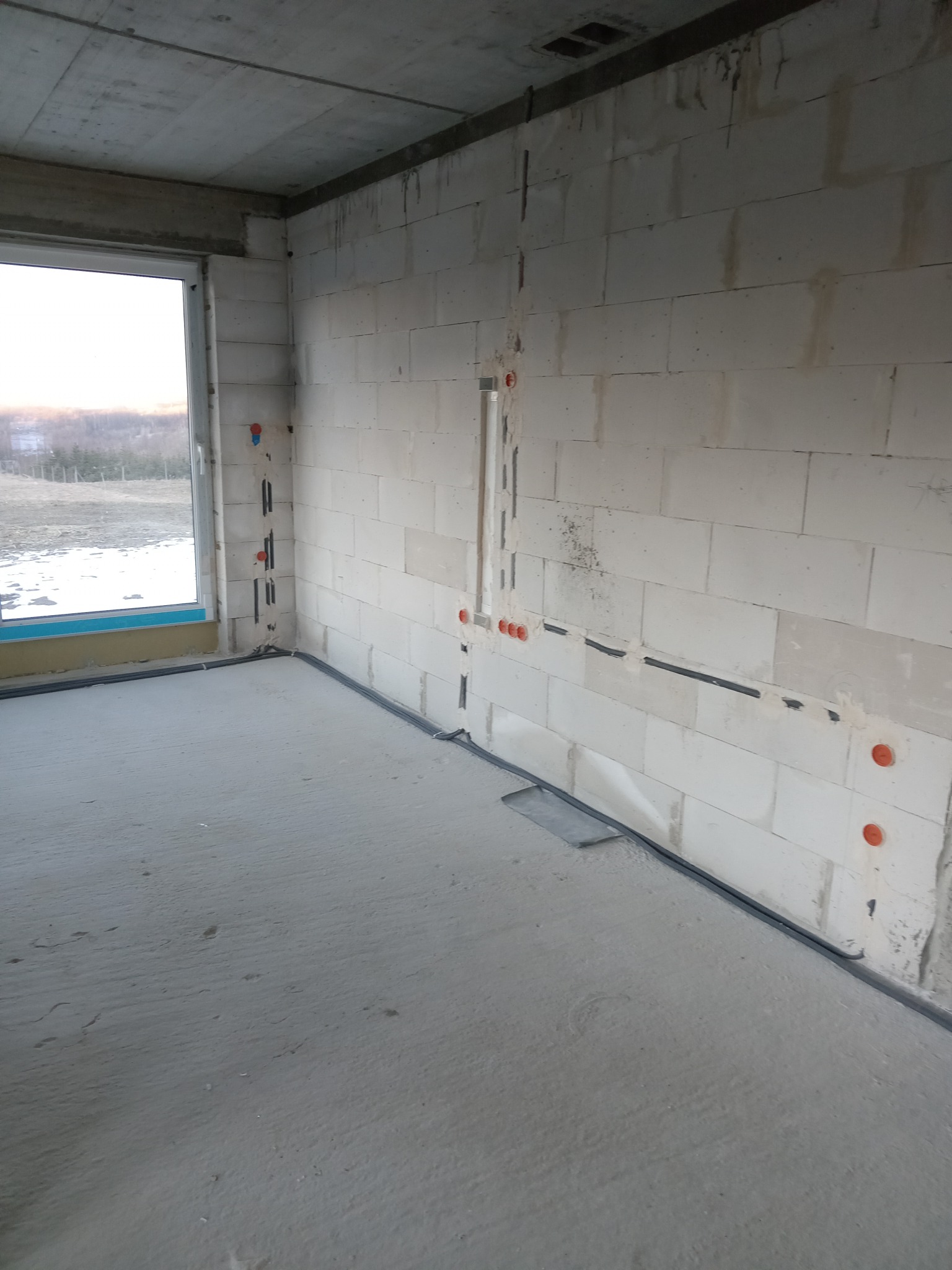 Surowy stan wnętrza z widoczną instalacją elektryczną w ścianach z betonu komórkowego i rurami przy podłodze. Okno z widokiem na zewnątrz.