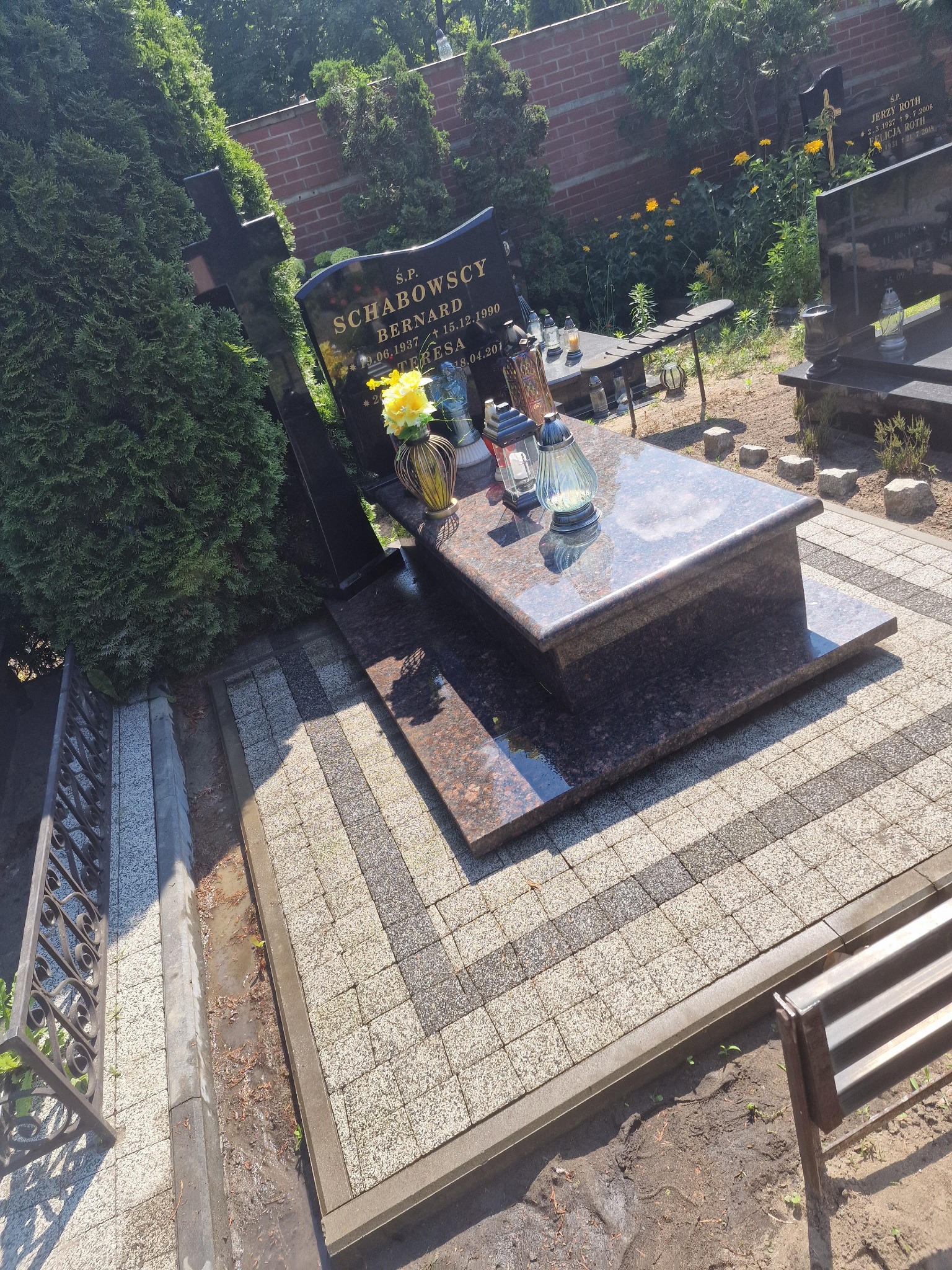 Granitowy nagrobek z kwiatami i zniczami na kostce brukowej w cieniu drzew, otoczony zielenią na cmentarzu, w słoneczny dzień.