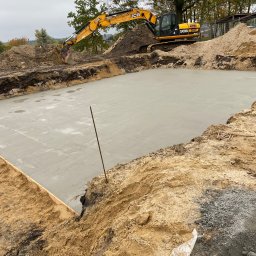 Oleksandr Kulyk - Świeżo wylana, gładka płyta betonowa fundamentowa na budowie z żółtą koparką JCB w tle. Widok z góry na wykop z deskowaniem i wystającą stalową prętem.