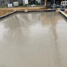 Świeżo wylana, mokra posadzka na budowie domu jednorodzinnego. W tle żółta betoniarka i biały budynek. Widoczne ślady po rozprowadzaniu betonu.