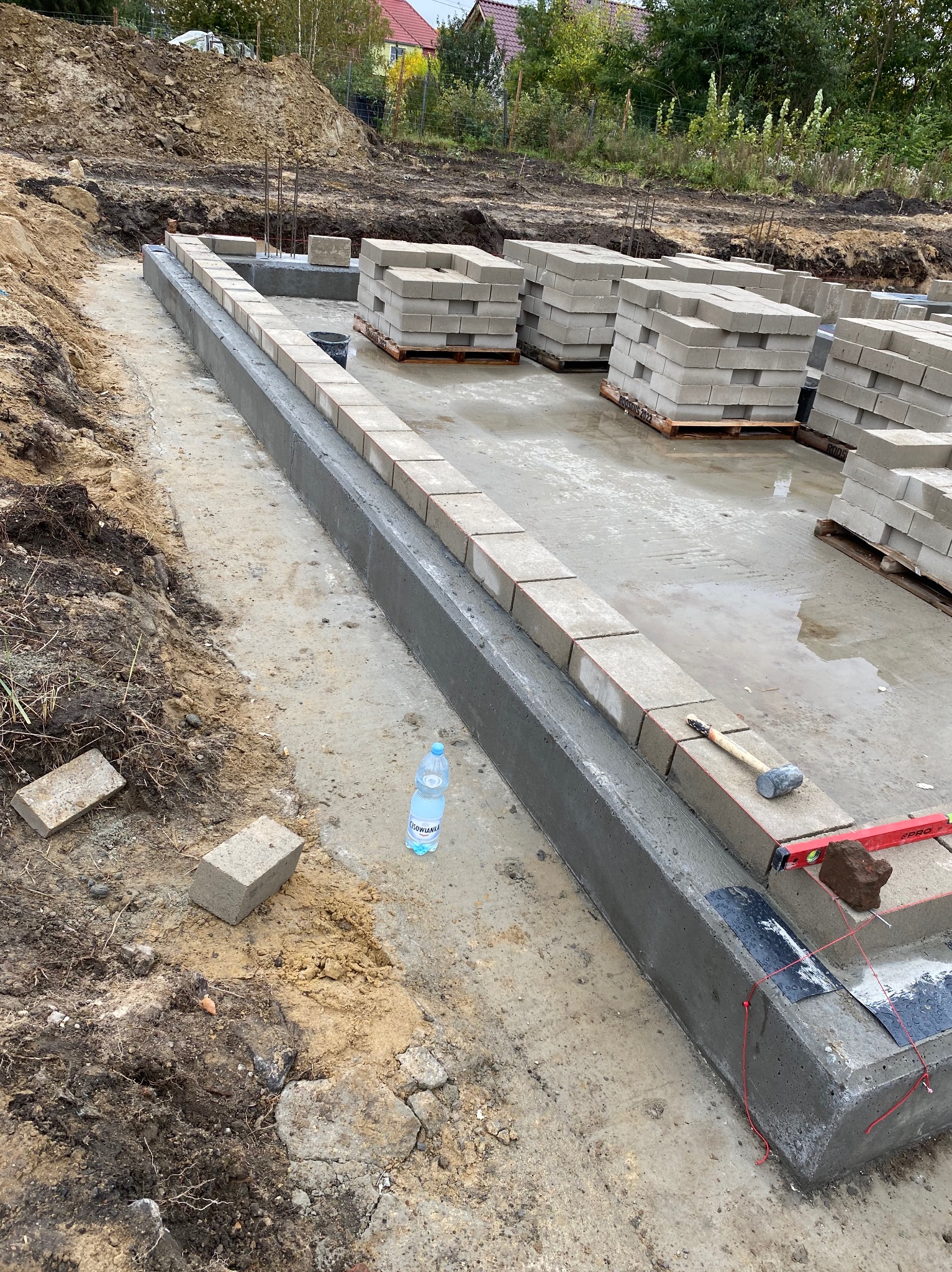 Fundamenty domu w trakcie budowy: zalane betonem ławy fundamentowe, ustawione bloczki betonowe, poziomica, młotek i butelka wody. Widok z góry.