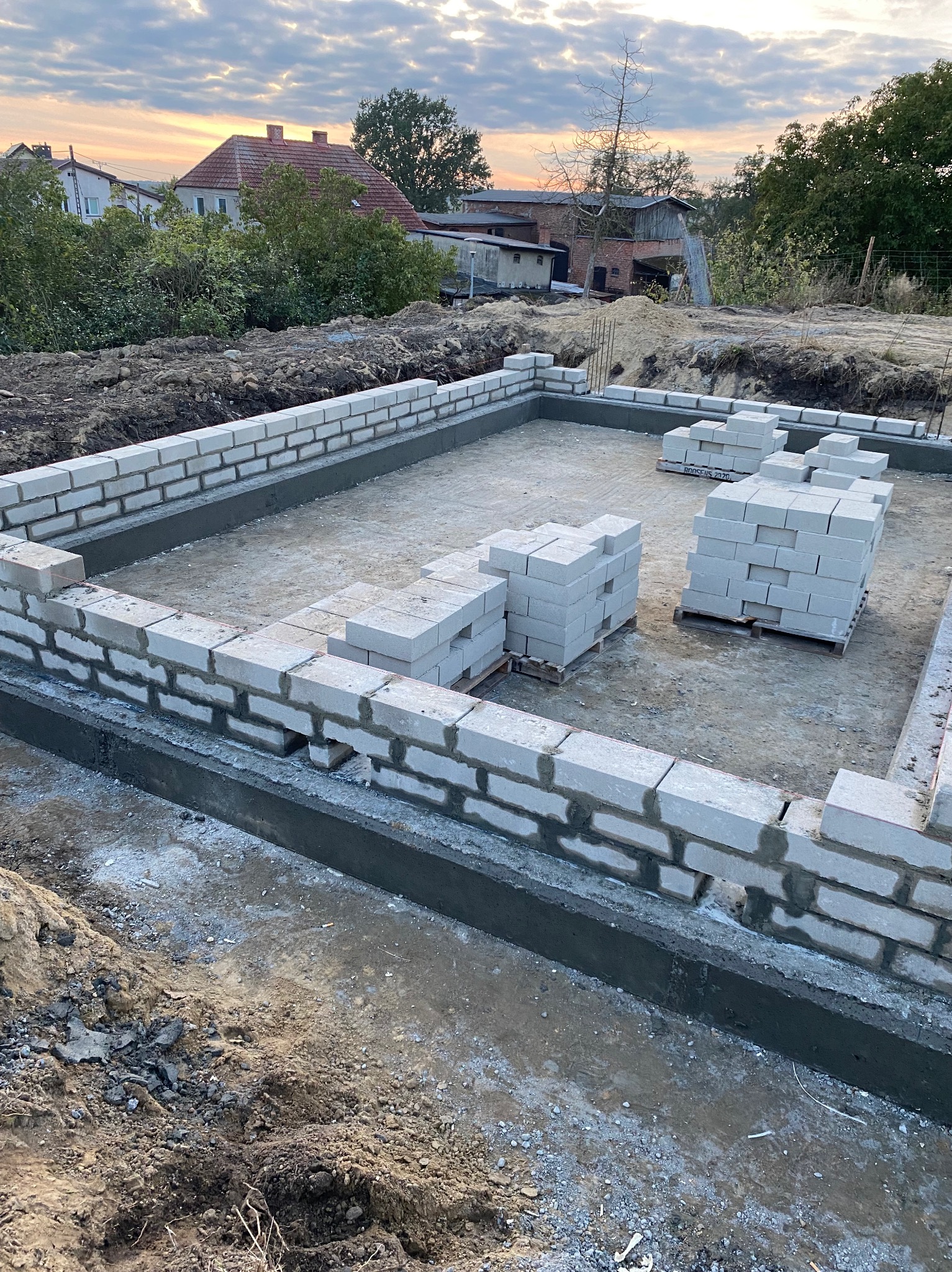 Wykonywanie fundamentów z bloczków betonowych, widoczne palety z bloczkami. Częściowo wzniesione ściany fundamentowe na tle zabudowy jednorodzinnej i zachodzącego słońca.