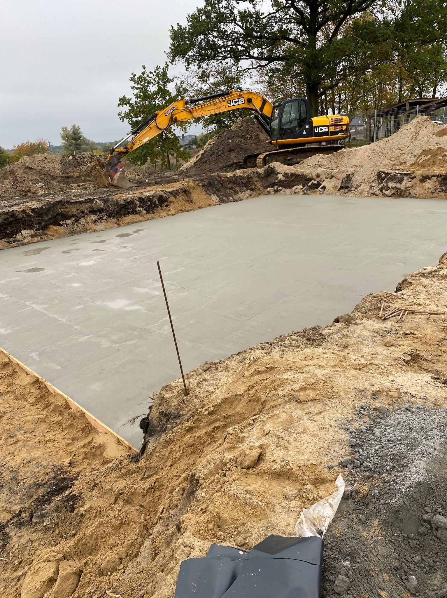 Świeżo wylana, gładka płyta betonowa fundamentowa na budowie z żółtą koparką JCB w tle. Widok z góry na wykop z deskowaniem i wystającą stalową prętem.