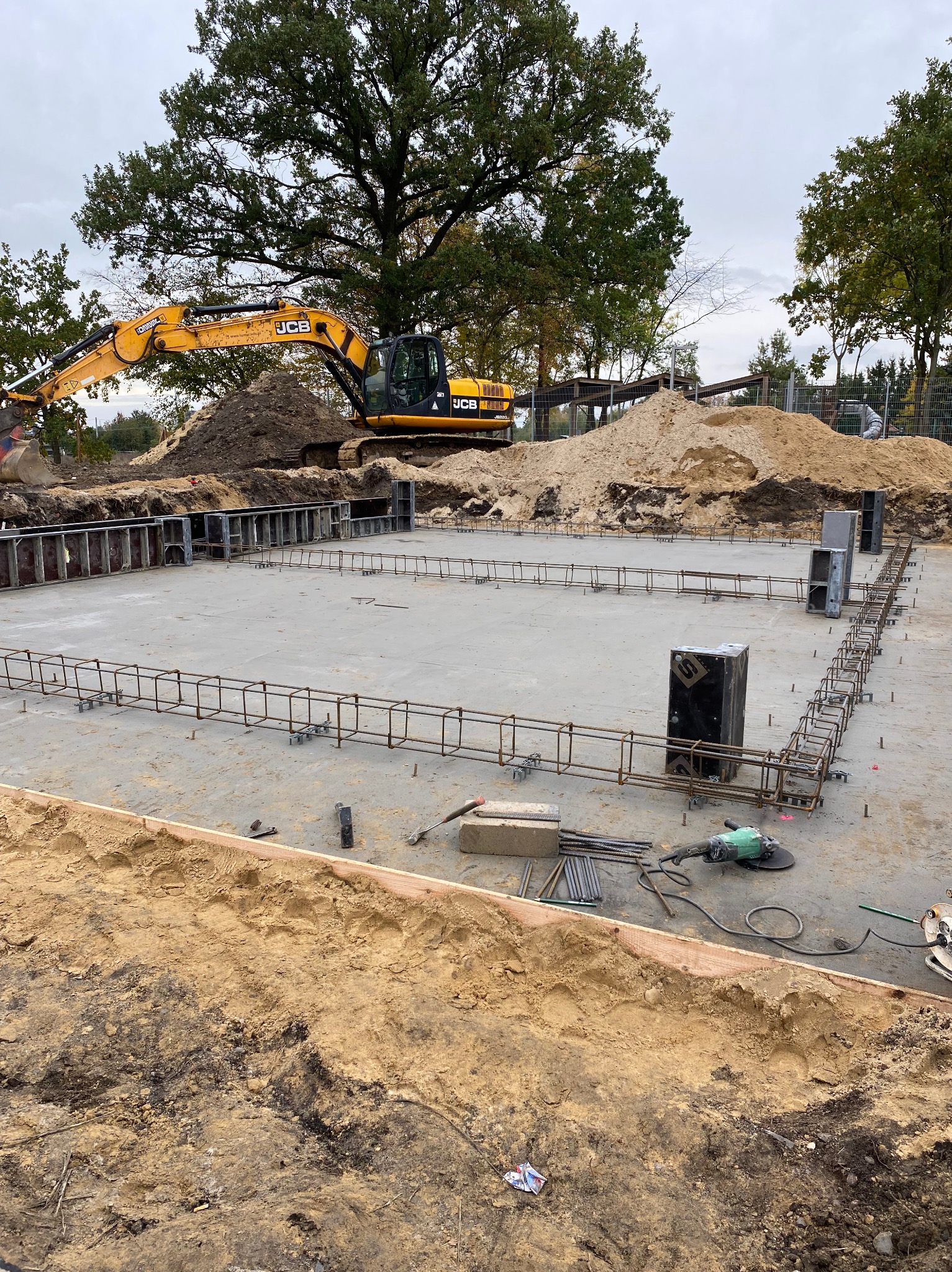 Betonowa płyta fundamentowa ze zbrojeniem, szalunki i koparka JCB w tle. Widoczne narzędzia na placu budowy. Fundamenty domu jednorodzinnego.