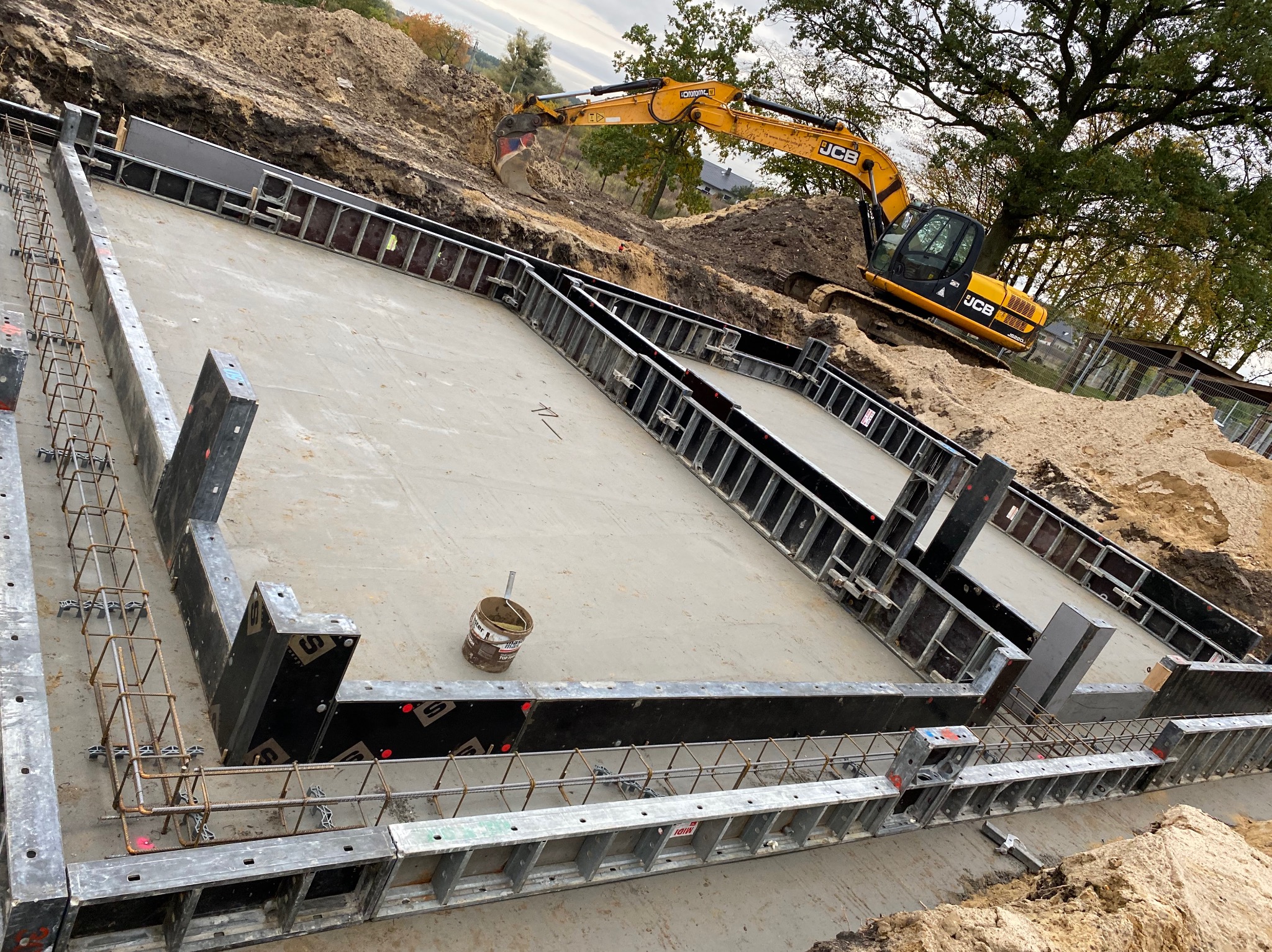 Wylany fundament z szalunkiem i zbrojeniem, na tle koparka JCB podczas prac ziemnych. Widok z góry na plac budowy.