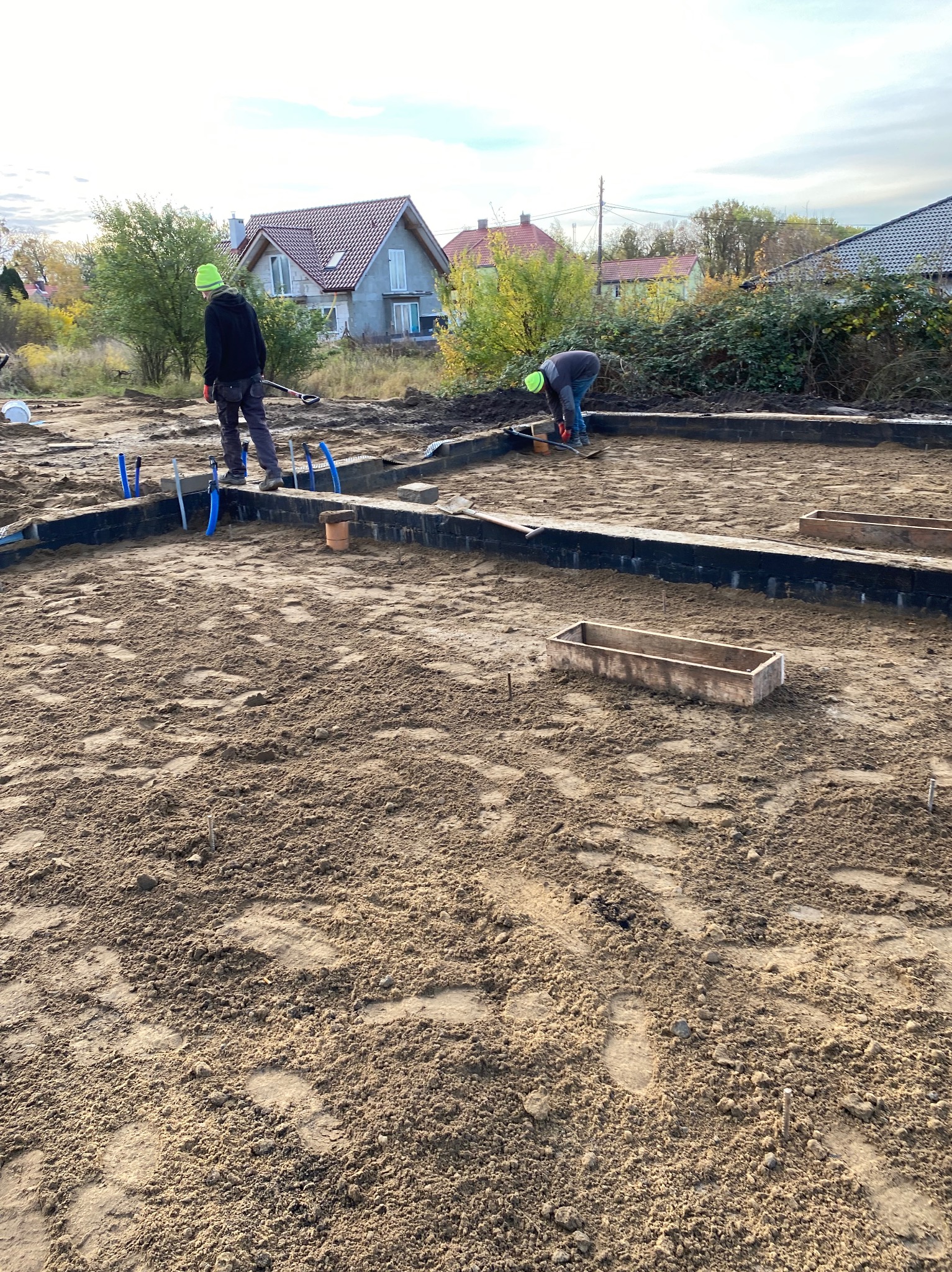 Budowa fundamentów: Dwóch pracowników w kaskach przygotowuje teren pod wylewkę, widoczne szalunki i wystające rury instalacyjne. W tle domy jednorodzinne.