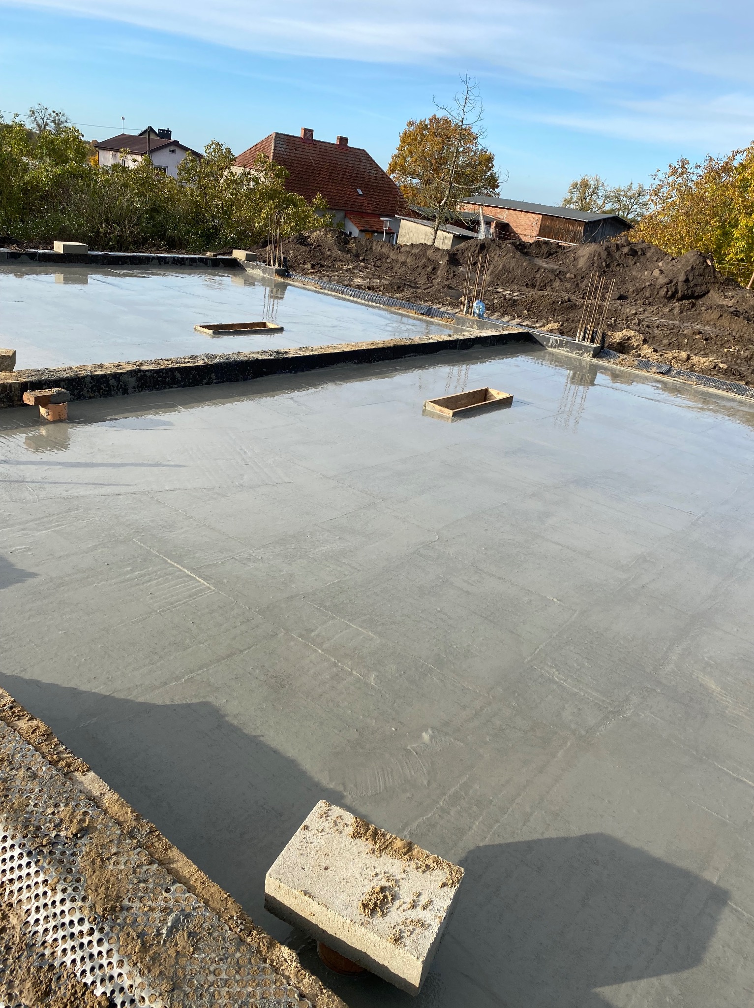 Świeżo wylana, gładka wylewka betonowa na budowie domu jednorodzinnego. Widoczne detale szalunków i otaczający teren budowy z ziemią i roślinnością.