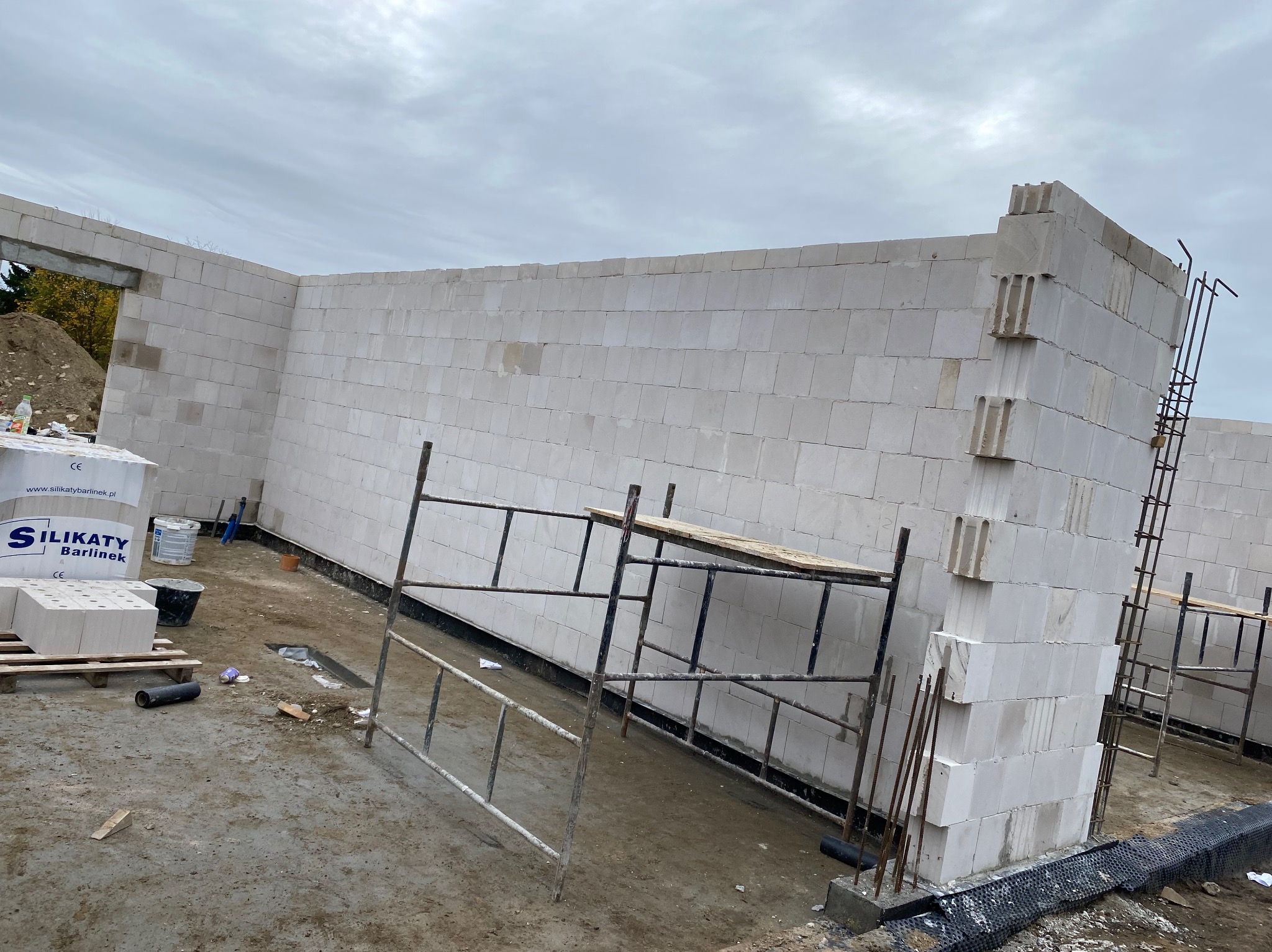 Wznoszenie ścian budynku z bloczków silikatowych. Widoczne rusztowania, paleta z bloczkami i częściowo ukończona konstrukcja. Szare niebo w tle.
