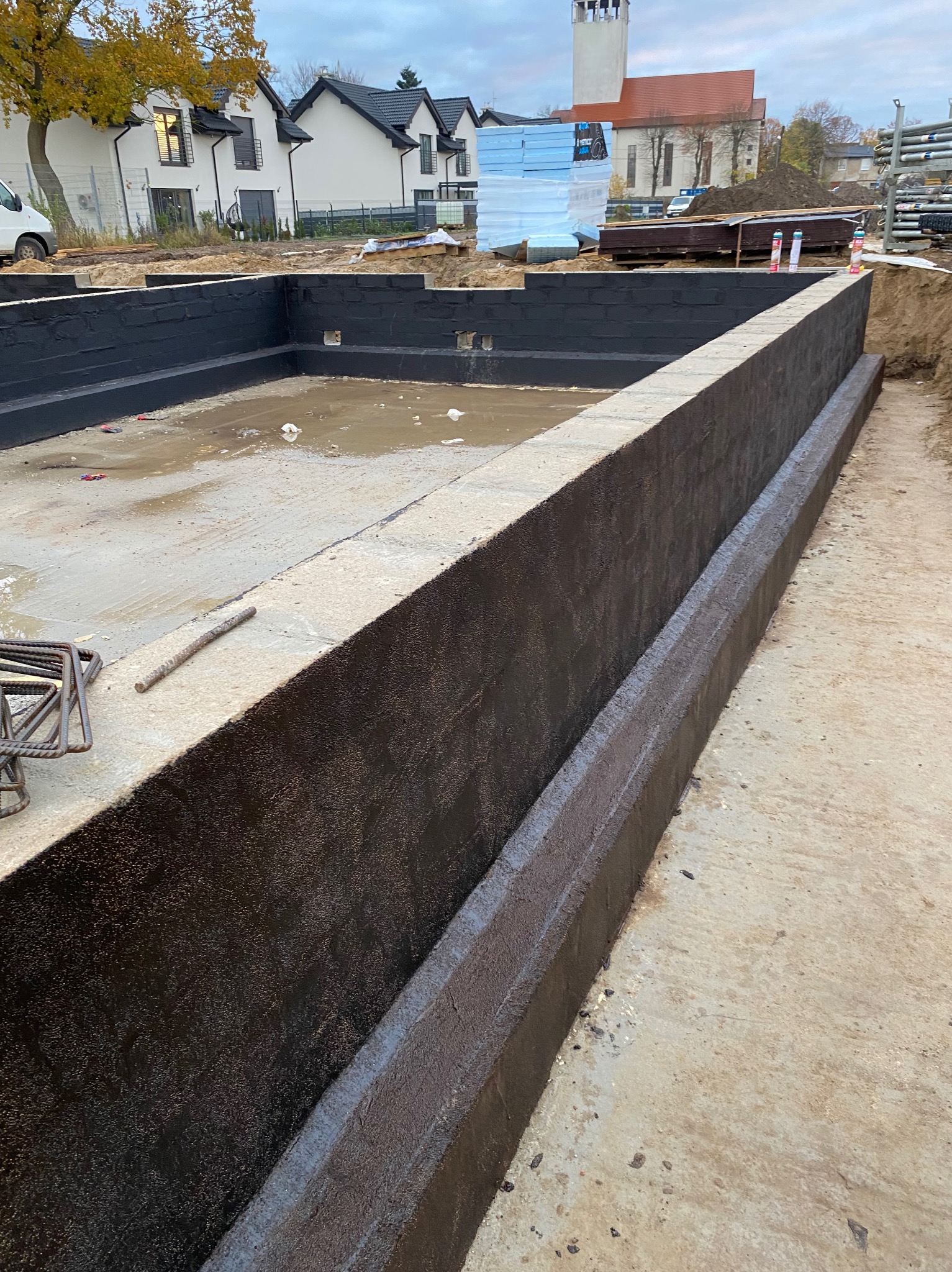 Zabezpieczone hydroizolacją ściany fundamentowe, widoczne zbrojenia i budynki w tle. Ujęcie z poziomu gruntu.