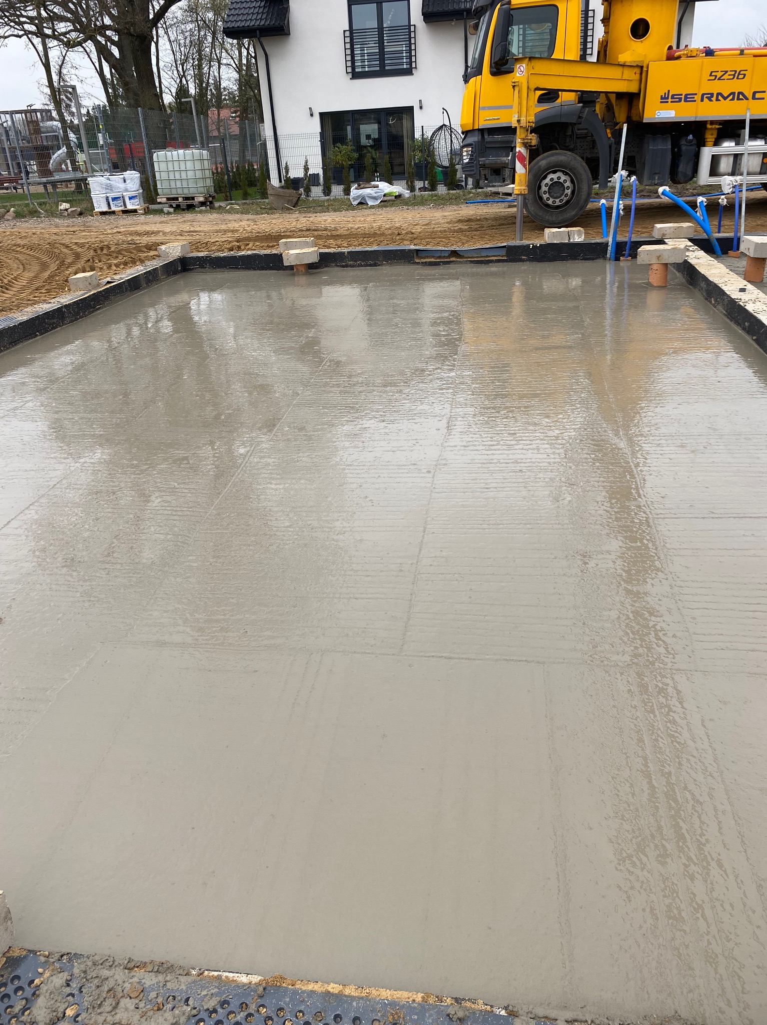 Świeżo wylana, mokra posadzka na budowie domu jednorodzinnego. W tle żółta betoniarka i biały budynek. Widoczne ślady po rozprowadzaniu betonu.