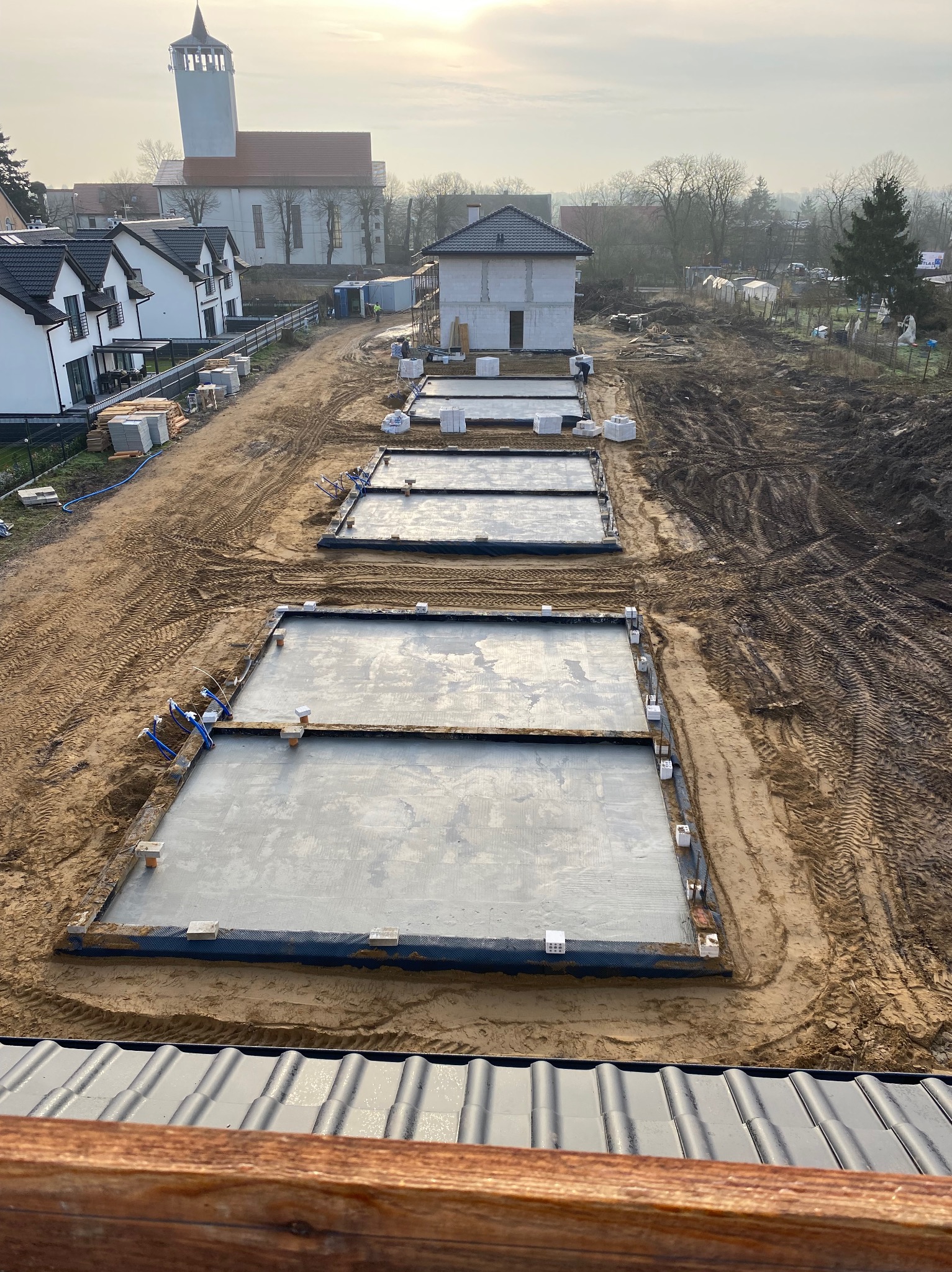 Wylane fundamenty pod budowę domów jednorodzinnych w krajobrazie z kościołem w tle. Widoczne przygotowane podłoże i instalacje.