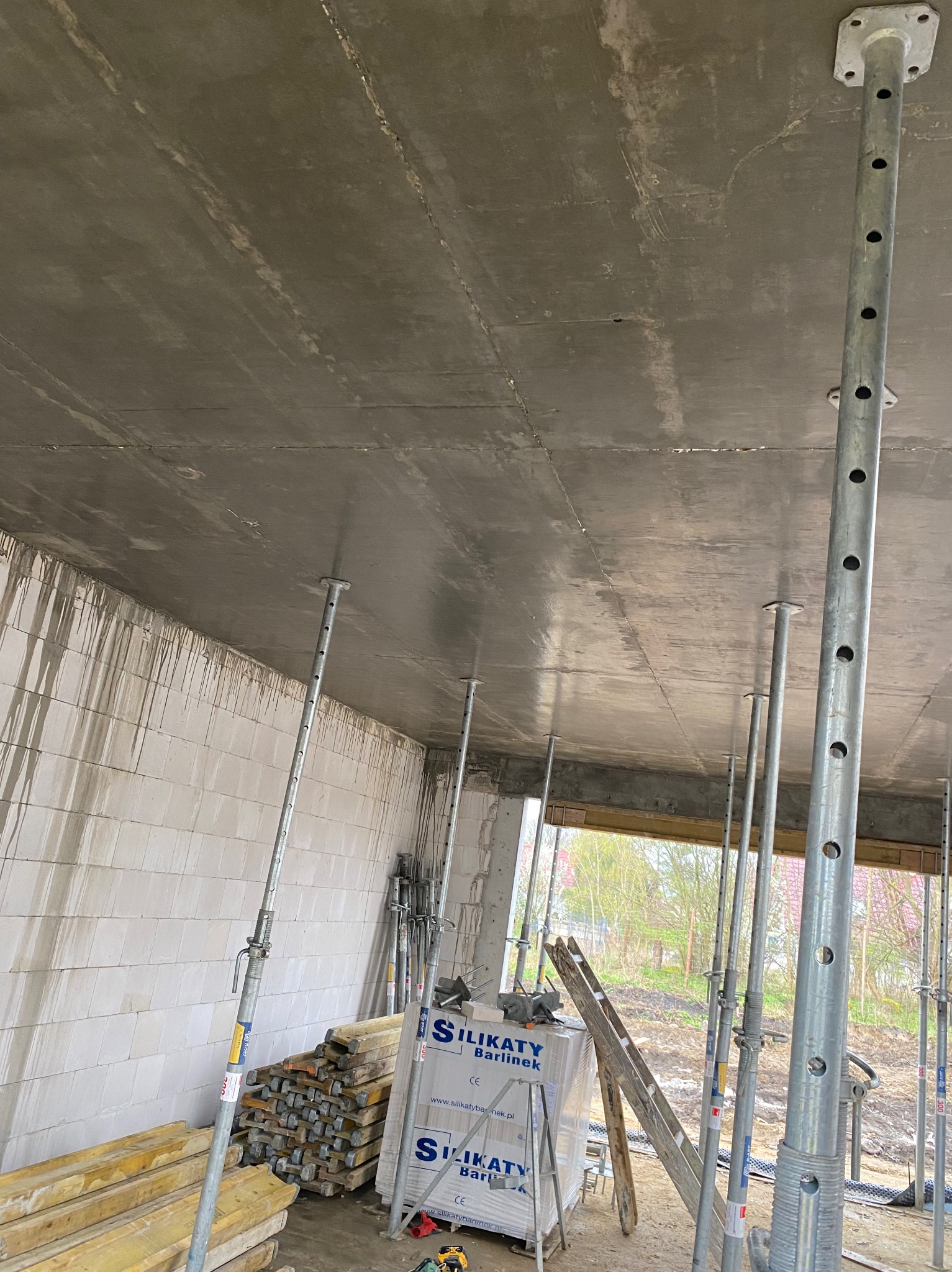 Świeżo wylany strop żelbetowy podparty metalowymi stemplami budowlanymi. Widoczne bloczki silikatowe i deski szalunkowe.