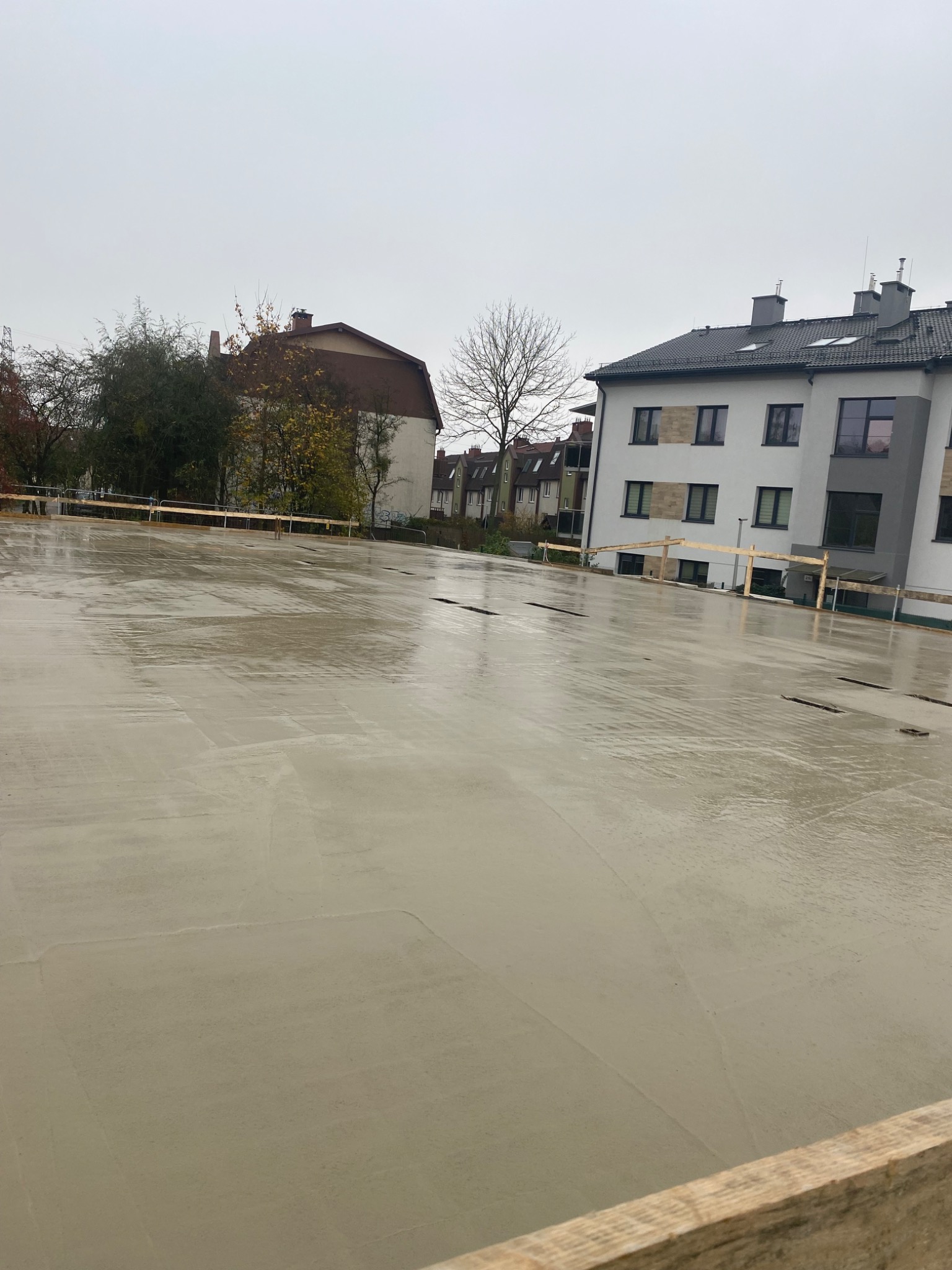 Świeżo wylana, mokra wylewka na budowie. Widoczne drewniane szalunki i fragmenty budynków w tle. Pochmurny dzień.