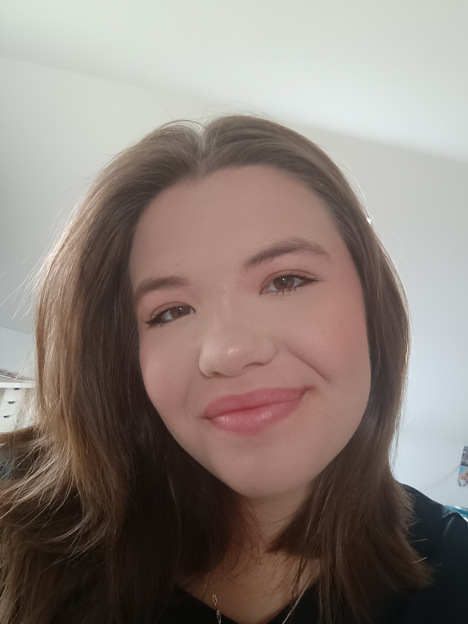 Portret uśmiechniętej brunetki z delikatnym makijażem, w ujęciu od dołu, z widocznym fragmentem garderoby i elementami wyposażenia wnętrza w tle.