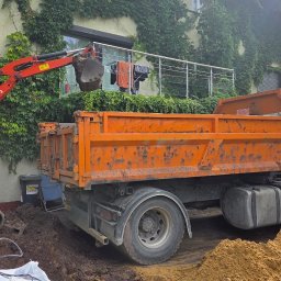 WEAR ELITE SPÓŁKA Z OGRANICZONĄ ODPOWIEDZIALNOŚCIĄ - Pomarańczowa wywrotka i minikoparka Kubota przy domu z bluszczem podczas prac ziemnych. Łyżka koparki nad skrzynią ładunkową. Widoczny operator w kabinie.