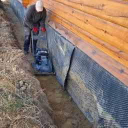 WEAR ELITE SPÓŁKA Z OGRANICZONĄ ODPOWIEDZIALNOŚCIĄ - Pracownik zagęszcza grunt w wąskim wykopie wzdłuż drewnianego domu, zabezpieczonego folią kubełkową. Widoczne narzędzie pracy i fragment trawnika.