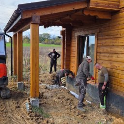 WEAR ELITE SPÓŁKA Z OGRANICZONĄ ODPOWIEDZIALNOŚCIĄ - Prace ziemne przy drewnianym domu: układanie drenażu z użyciem minikoparki Kubota. Widoczni pracownicy i przygotowany wykop.