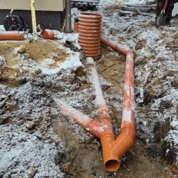 WEAR ELITE SPÓŁKA Z OGRANICZONĄ ODPOWIEDZIALNOŚCIĄ - Montaż pomarańczowych rur drenażowych w wykopie pokrytym śniegiem, widoczny pracownik z łopatą i budynek w tle. Zimowa sceneria.